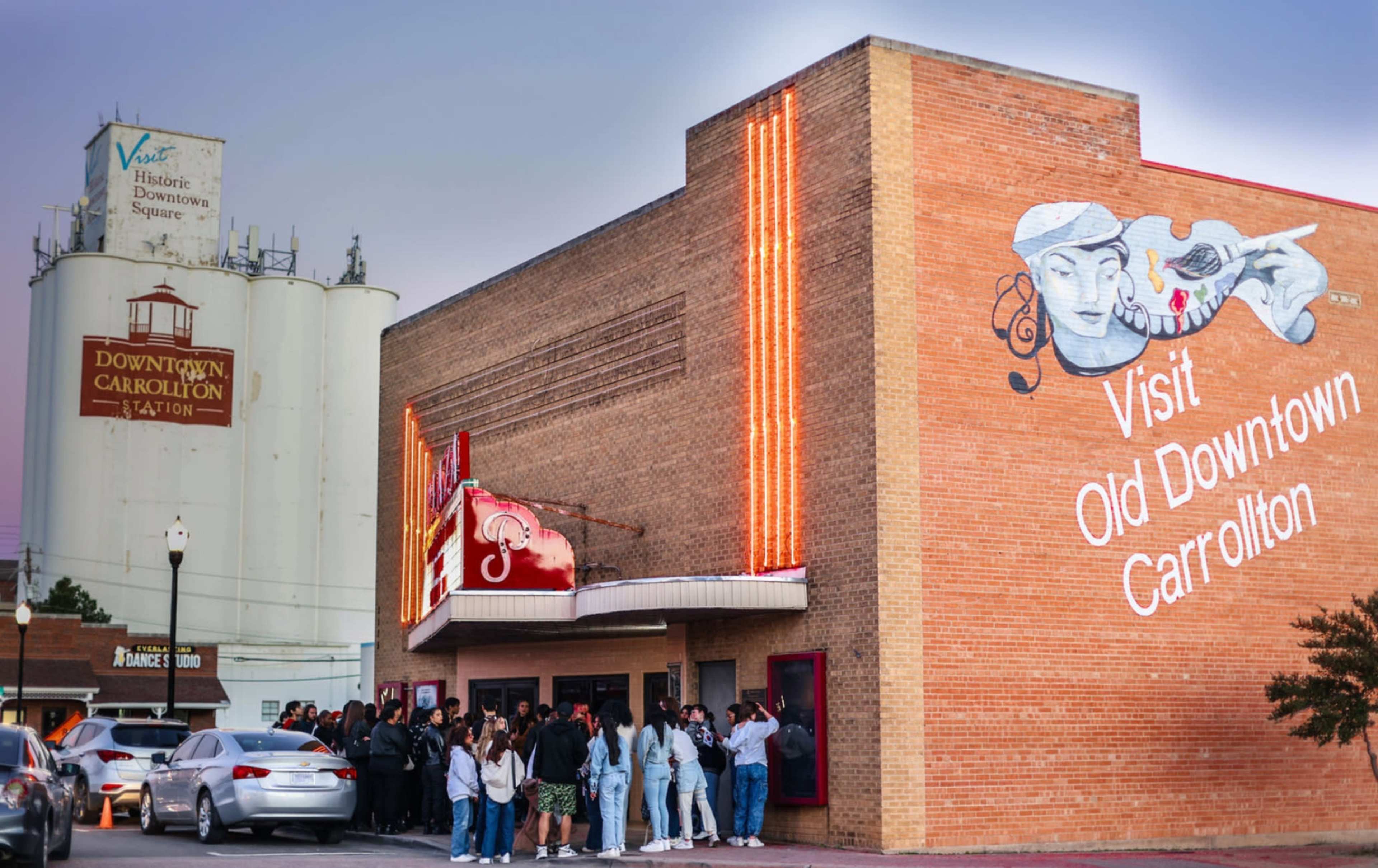 A crowd gathers outside a brick theater adorned with a colorful mural in downtown Carrollton.