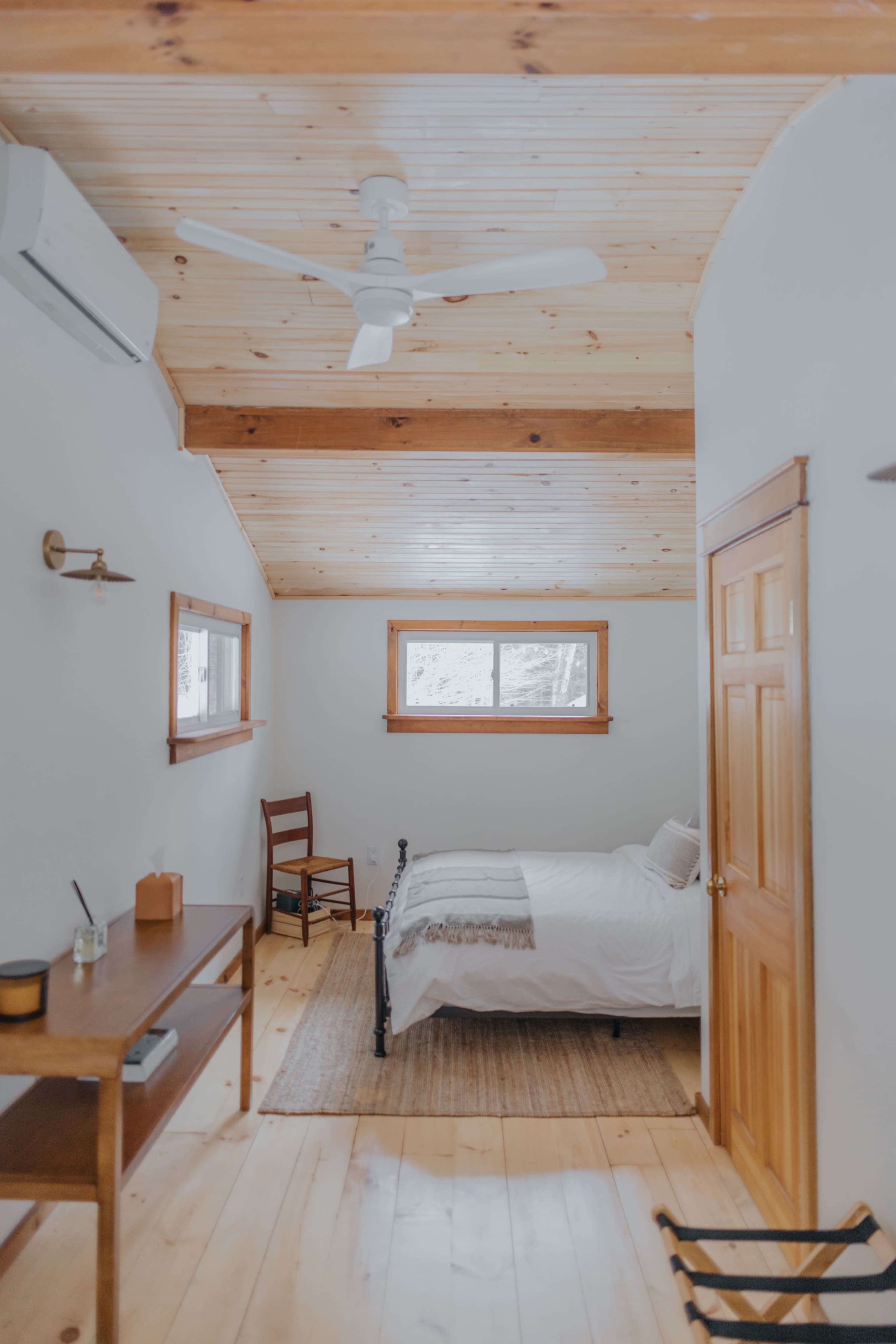 The image shows a simple bedroom with a bed, a wooden table, a chair, and a ceiling fan, all arranged under a wooden-paneled ceiling.
