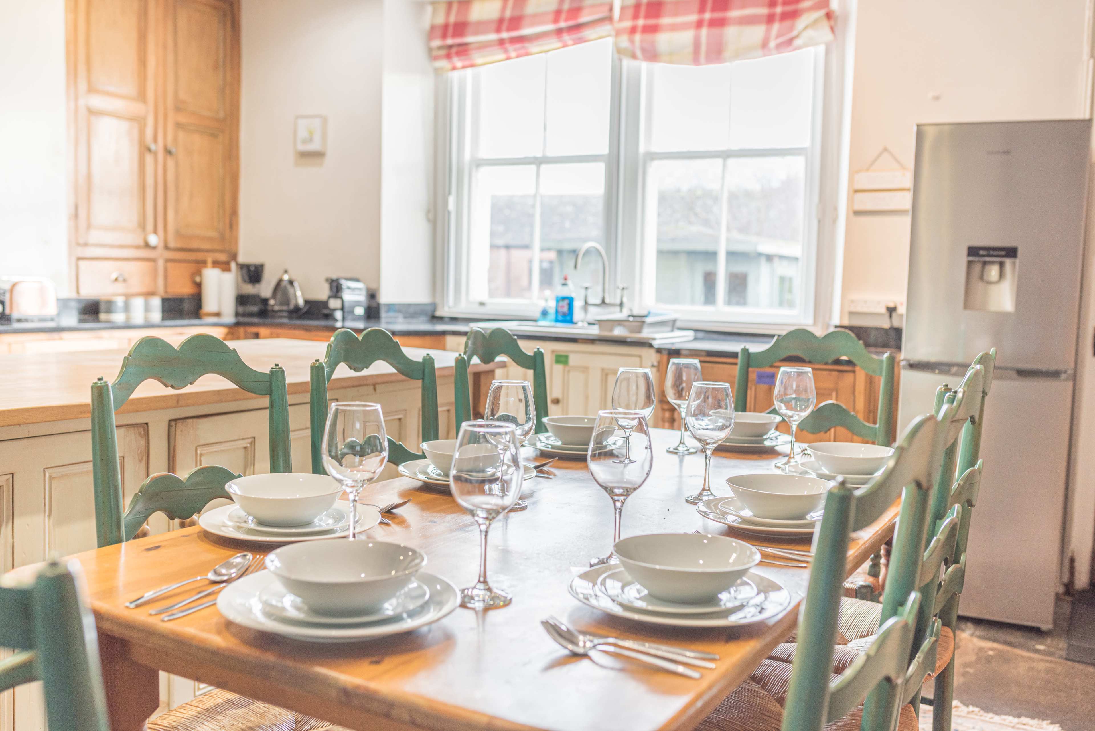 A dining table is set with plates and wine glasses in a kitchen with wooden cabinets and a window featuring a checkered curtain.