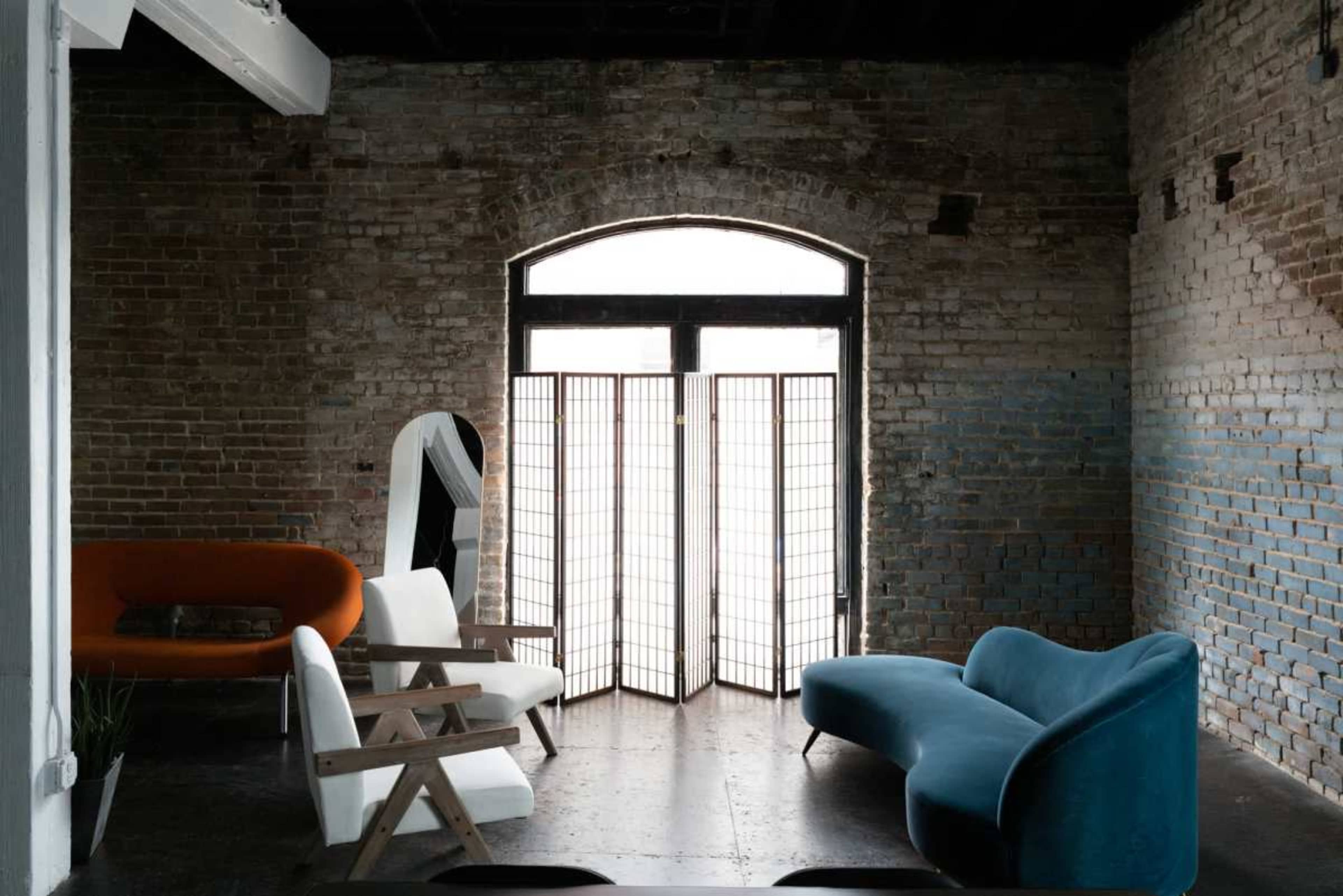 The image depicts a minimalist interior space featuring a mix of modern furniture, including a blue curved sofa, a white armchair, and an orange upholstered seat, alongside a folding screen in front of a large window.