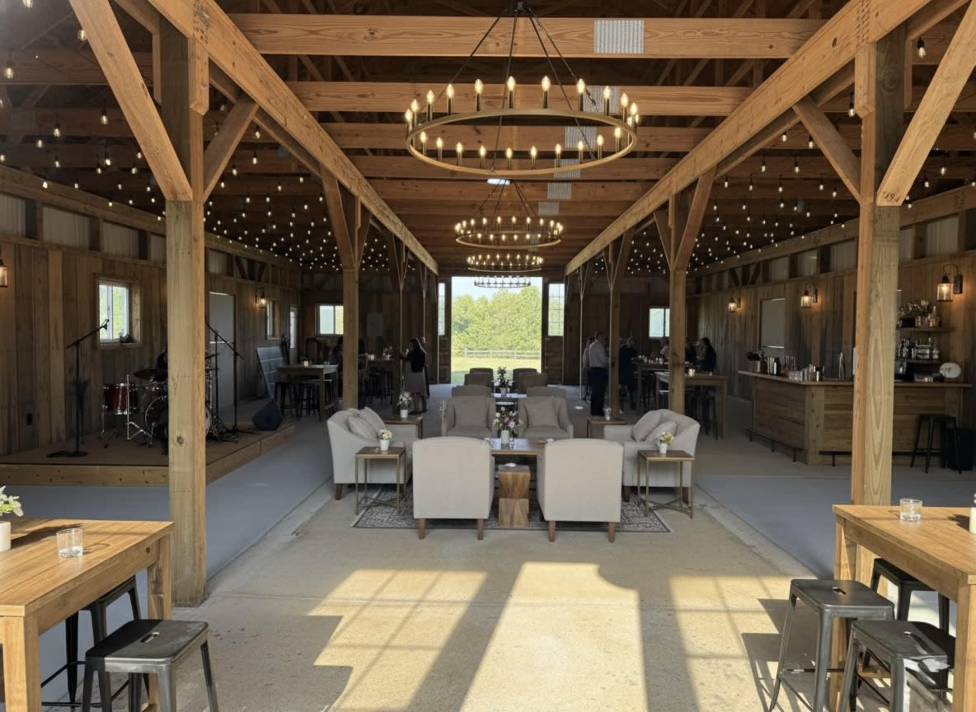 The image shows a spacious wooden interior of an event venue with tables, chairs, and a small stage area, illuminated by hanging lights.
