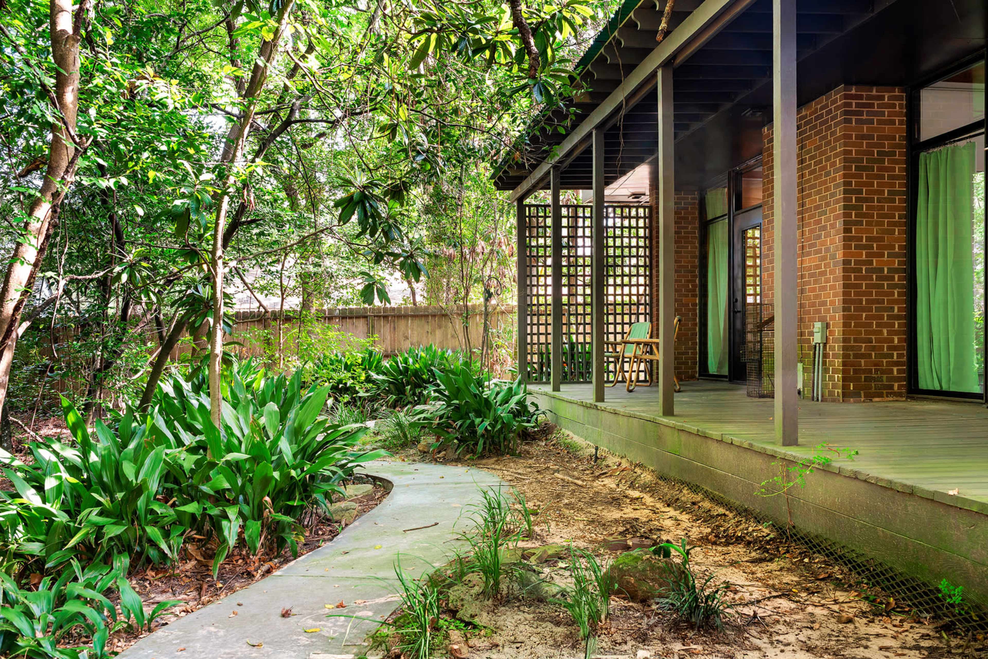 A pathway winds through lush greenery leading to a modern brick house with large windows and a wooden deck.