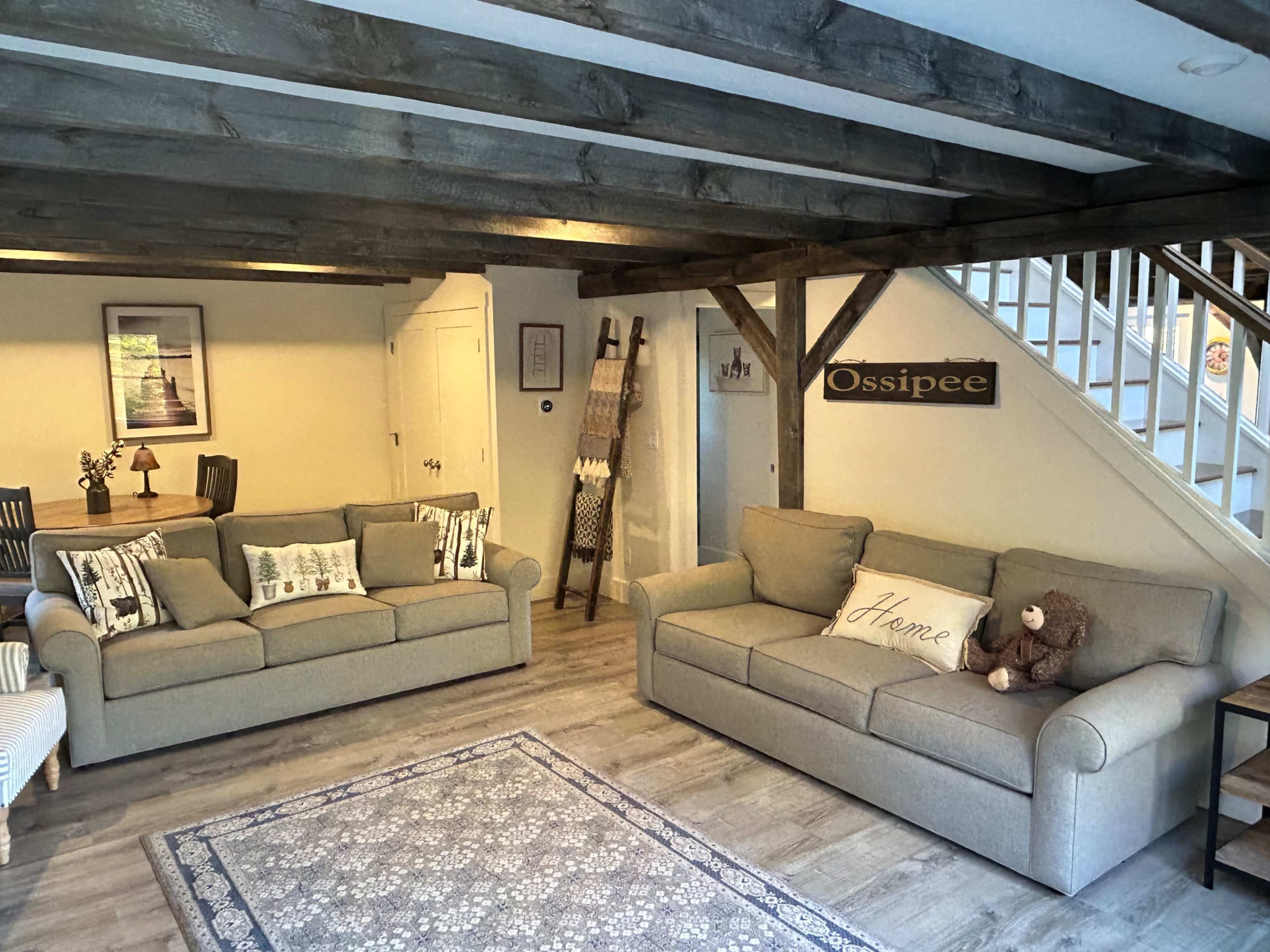 The living room features two beige sofas, a decorative rug, a wooden beam ceiling, and a staircase in the background.