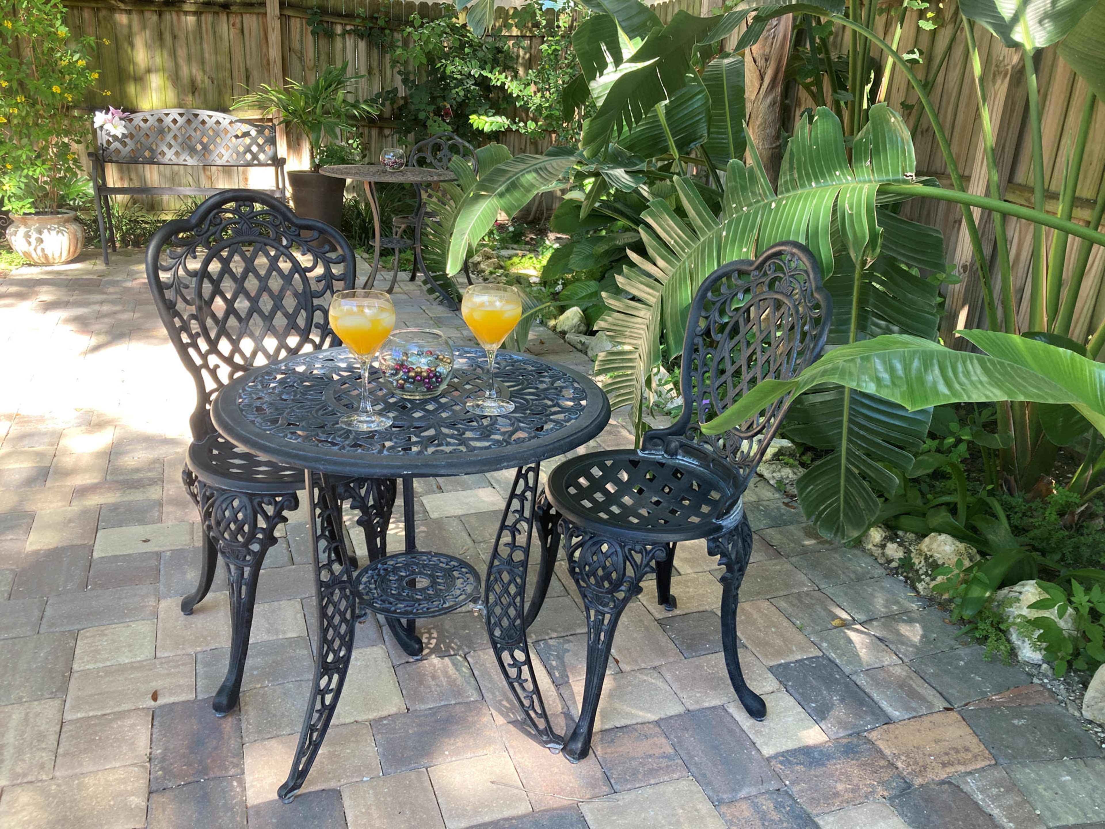 A wrought iron table with two chairs is set on a brick patio surrounded by lush green plants, with two glasses of orange beverages placed on the table.