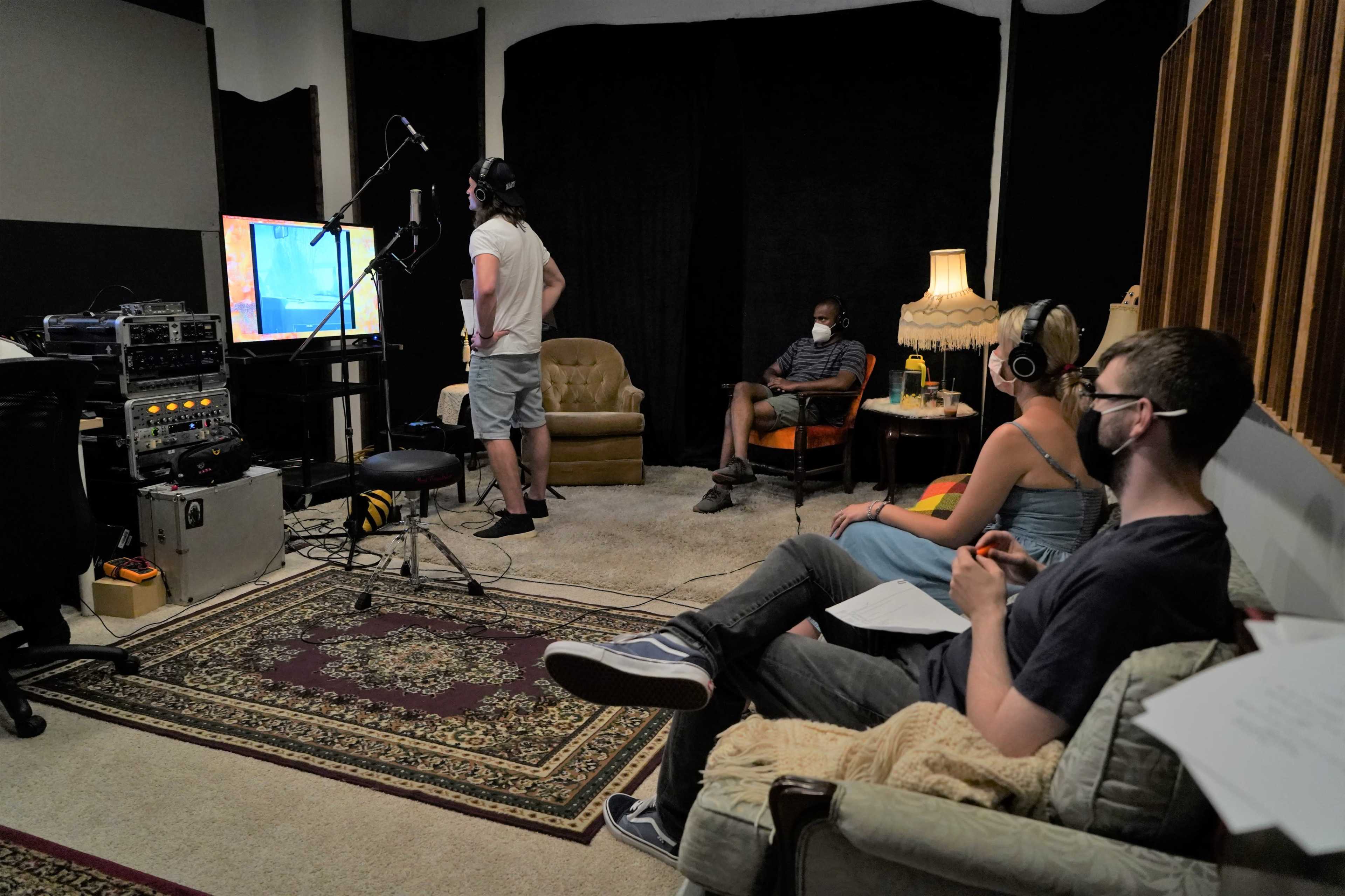 A group of four people is recording in a studio, with two seated and wearing headphones while a third stands near a microphone, watching a screen displaying animation.