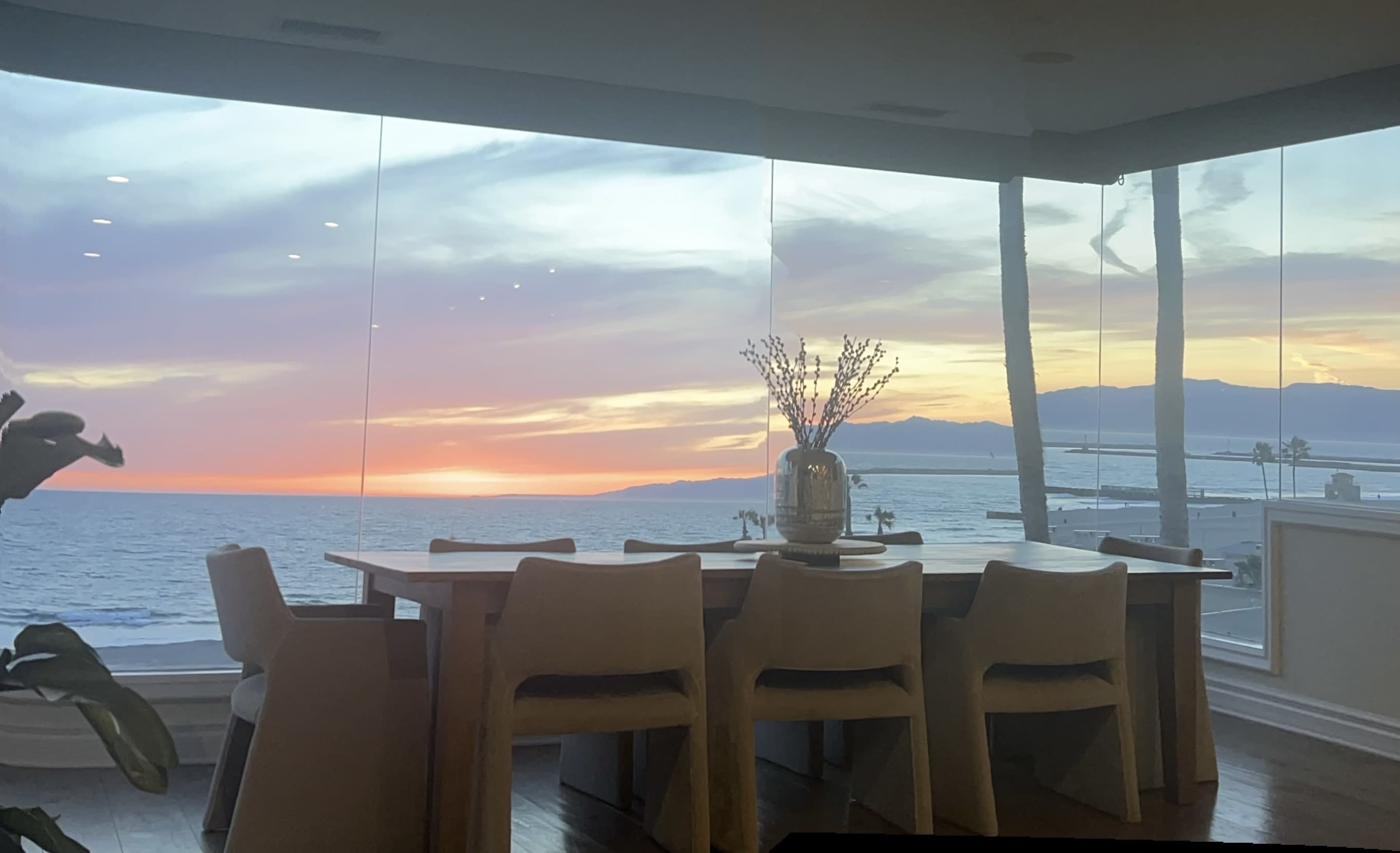 A dining area with a wooden table and chairs overlooks a sunset over the ocean and mountains through large glass windows.