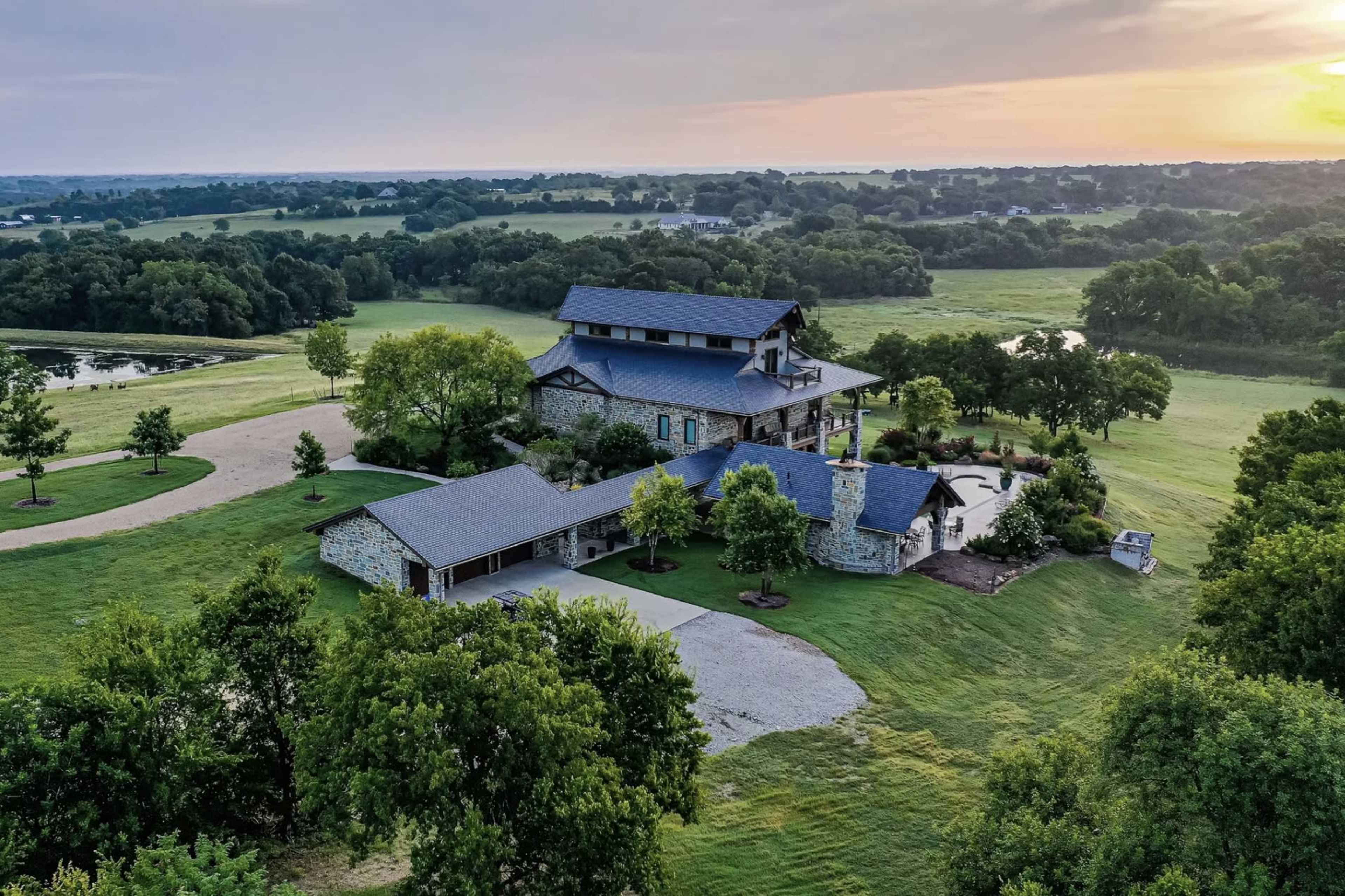 A large stone house with multiple roofs is surrounded by lush green fields and trees under a colorful sunset.