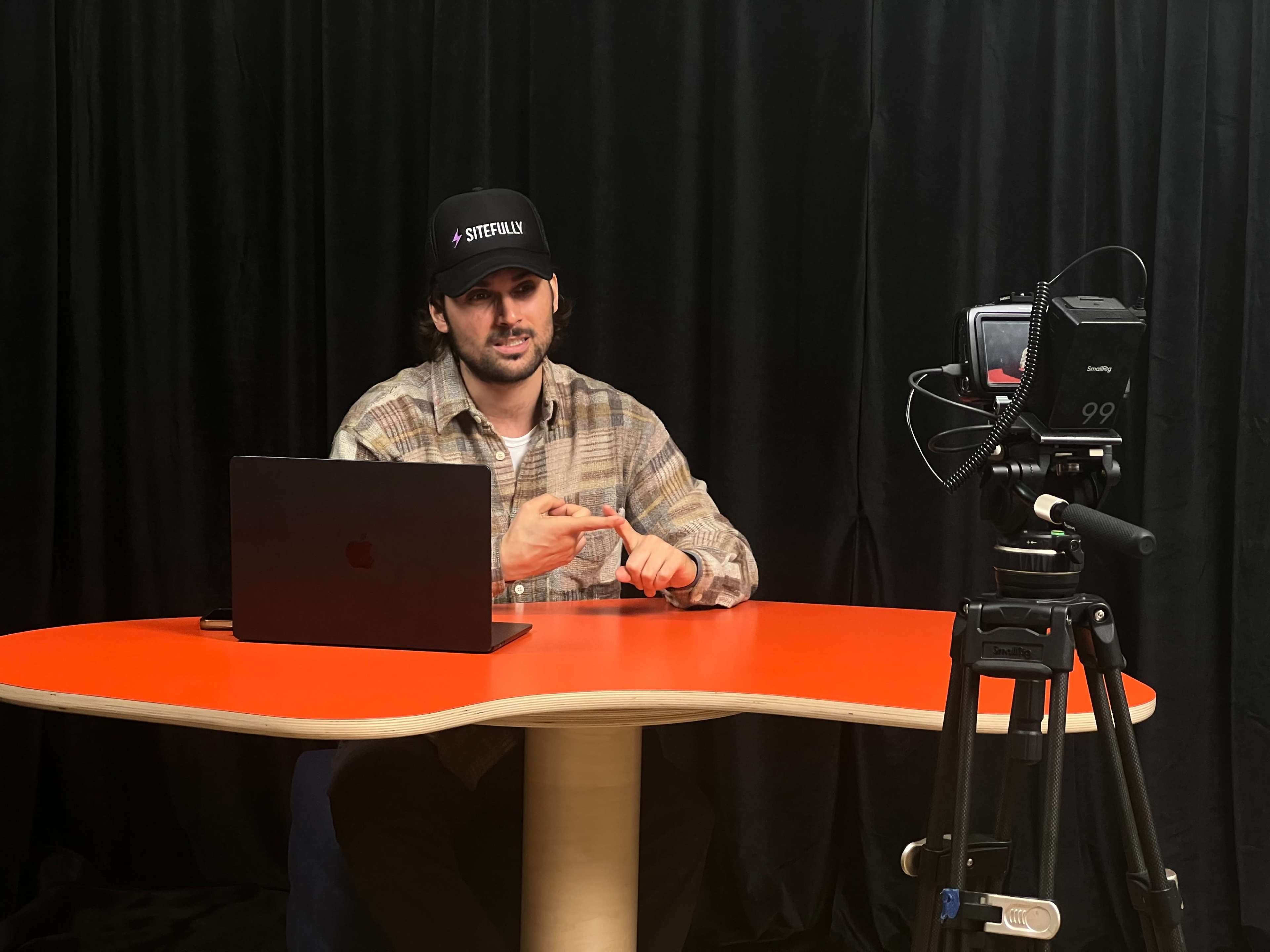 A man sits at a bright orange table, speaking into a camera while a laptop is open in front of him.