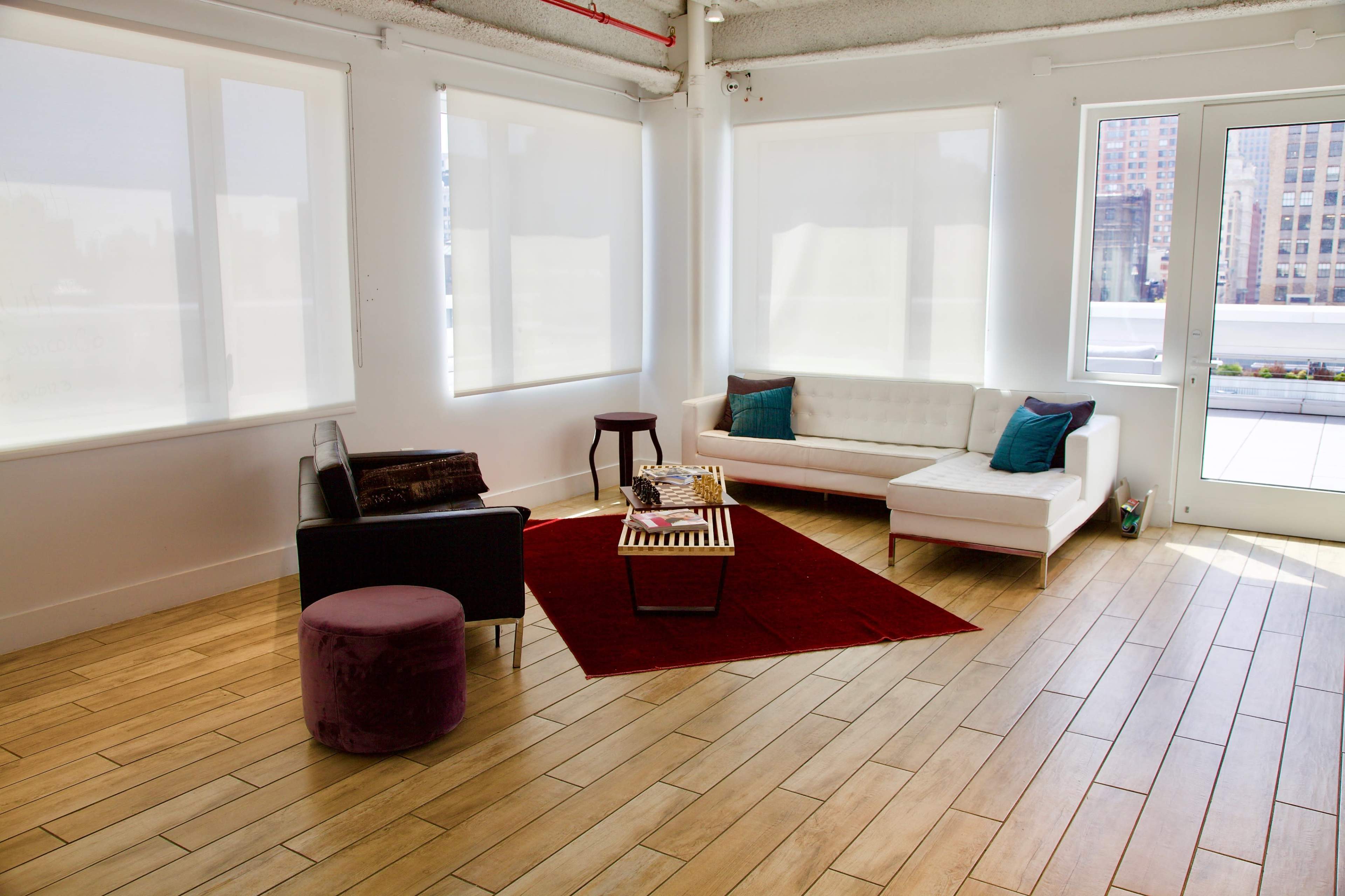 A bright, modern living space with a white sectional sofa, a black chair, a round purple ottoman, and a red area rug on wooden flooring.