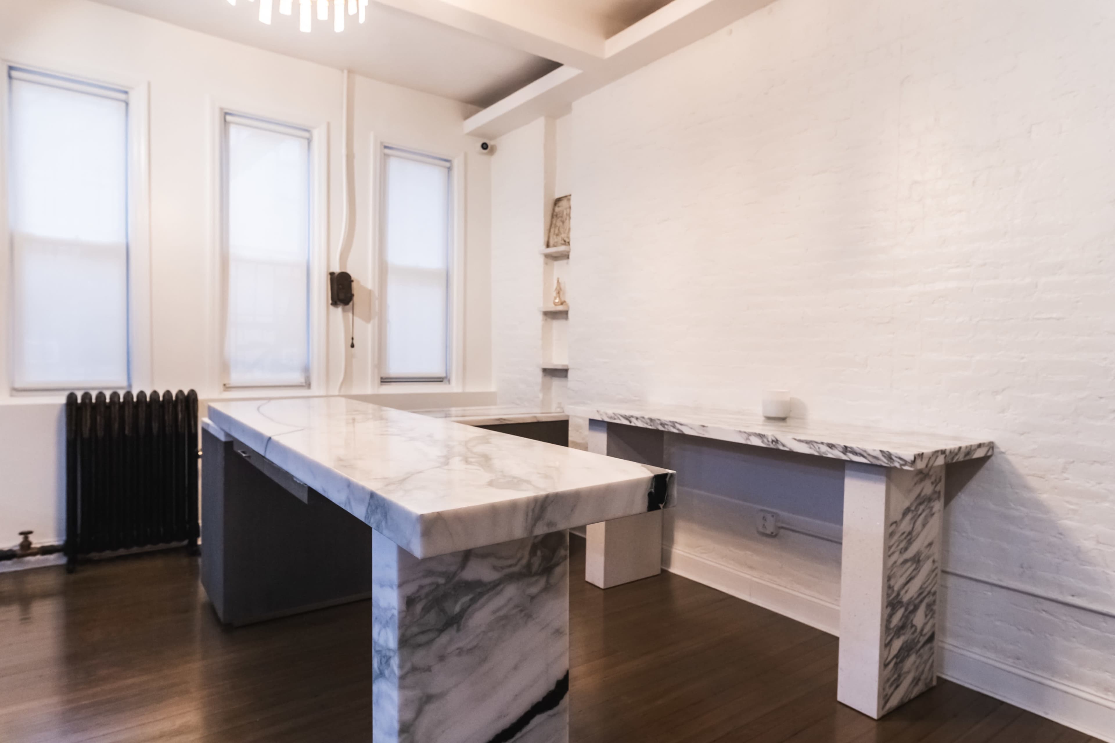 The image shows a spacious room with white brick walls, featuring a large marble table and a smaller marble console against the wall, with minimal furnishings and natural light from nearby windows.