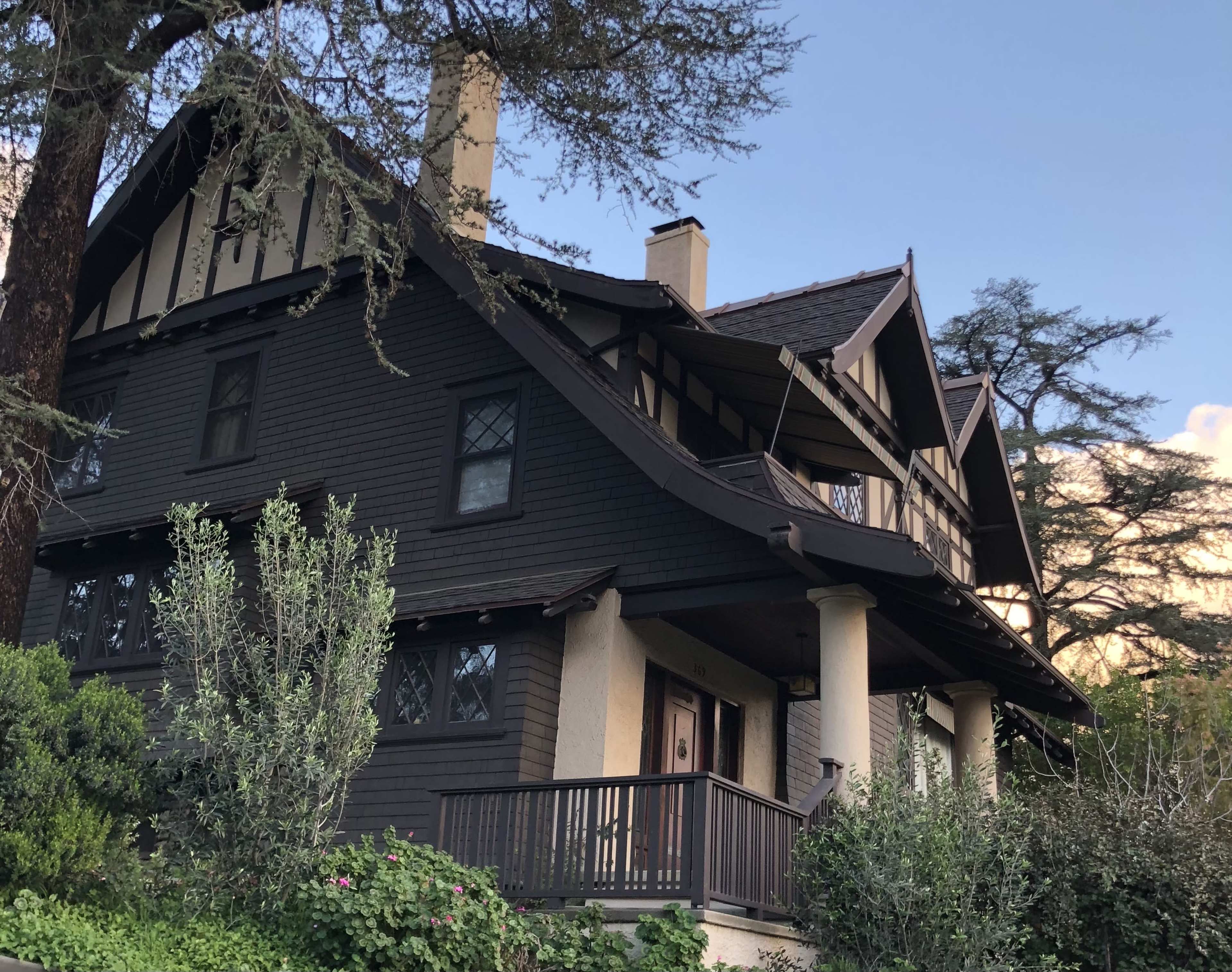 A large, dark wooden house with a steep roof, prominent gables, and a front porch surrounded by greenery.
