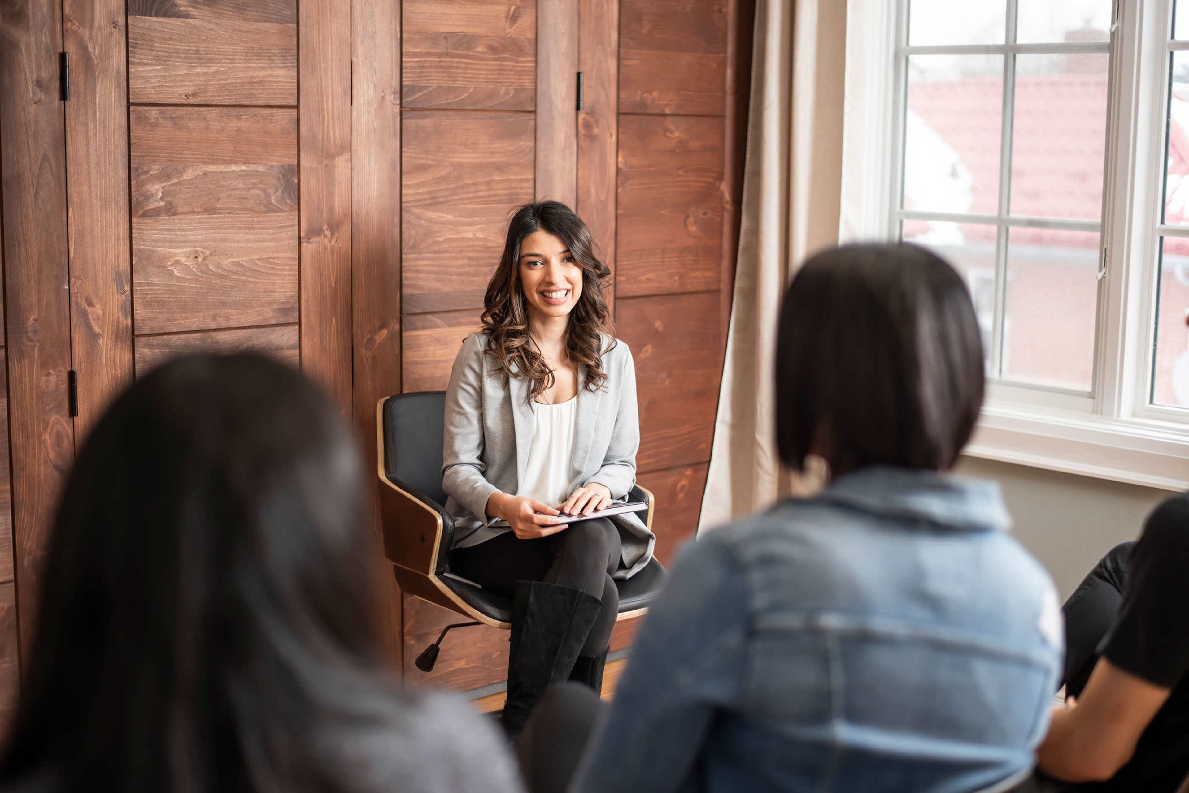 A woman is seated in a chair, smiling and engaging with a small group of people seated in front of her in a room with wooden walls and natural light.