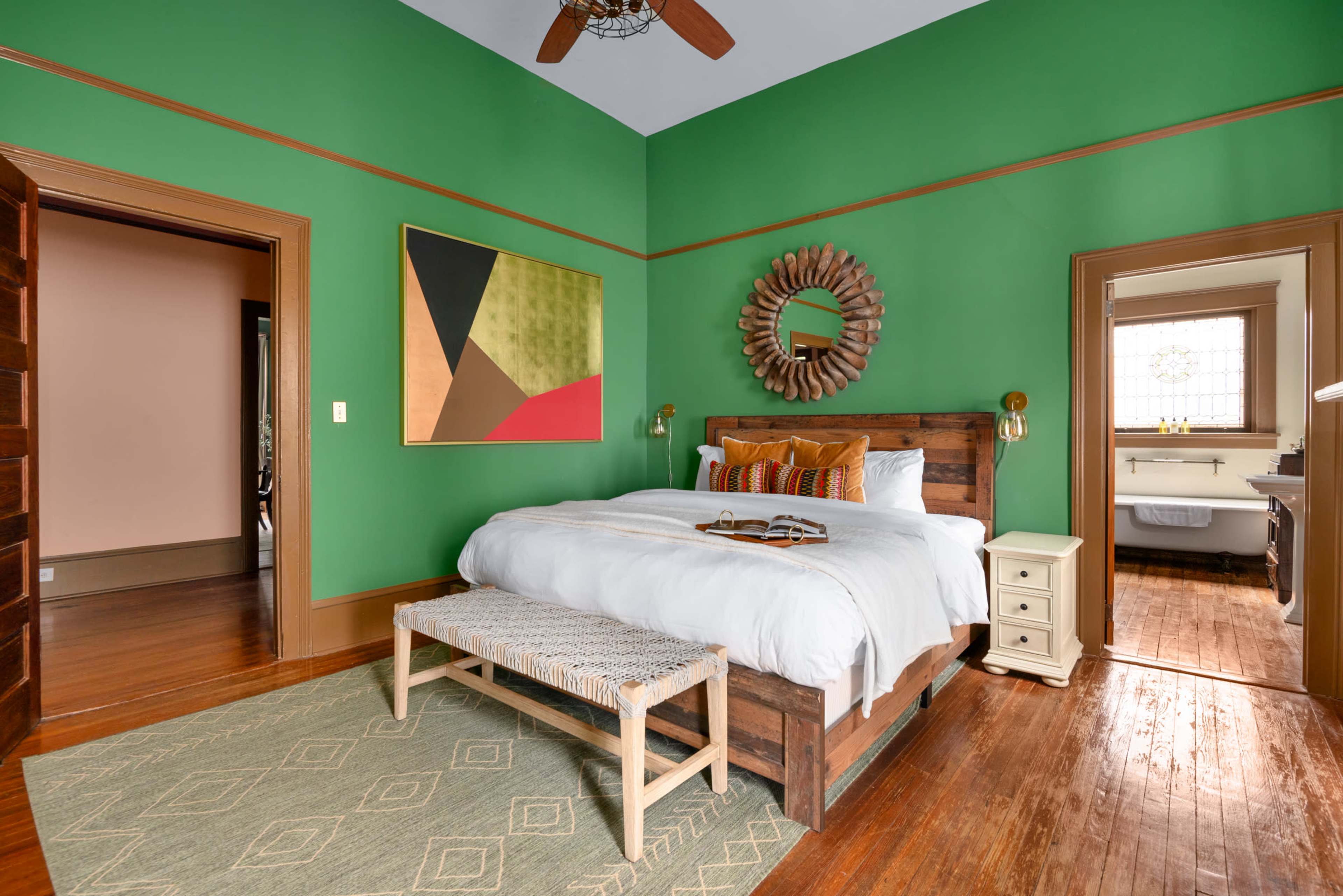 The room features a wooden bed with white bedding, a woven bench at the foot, green walls, and a large piece of abstract art above the headboard.