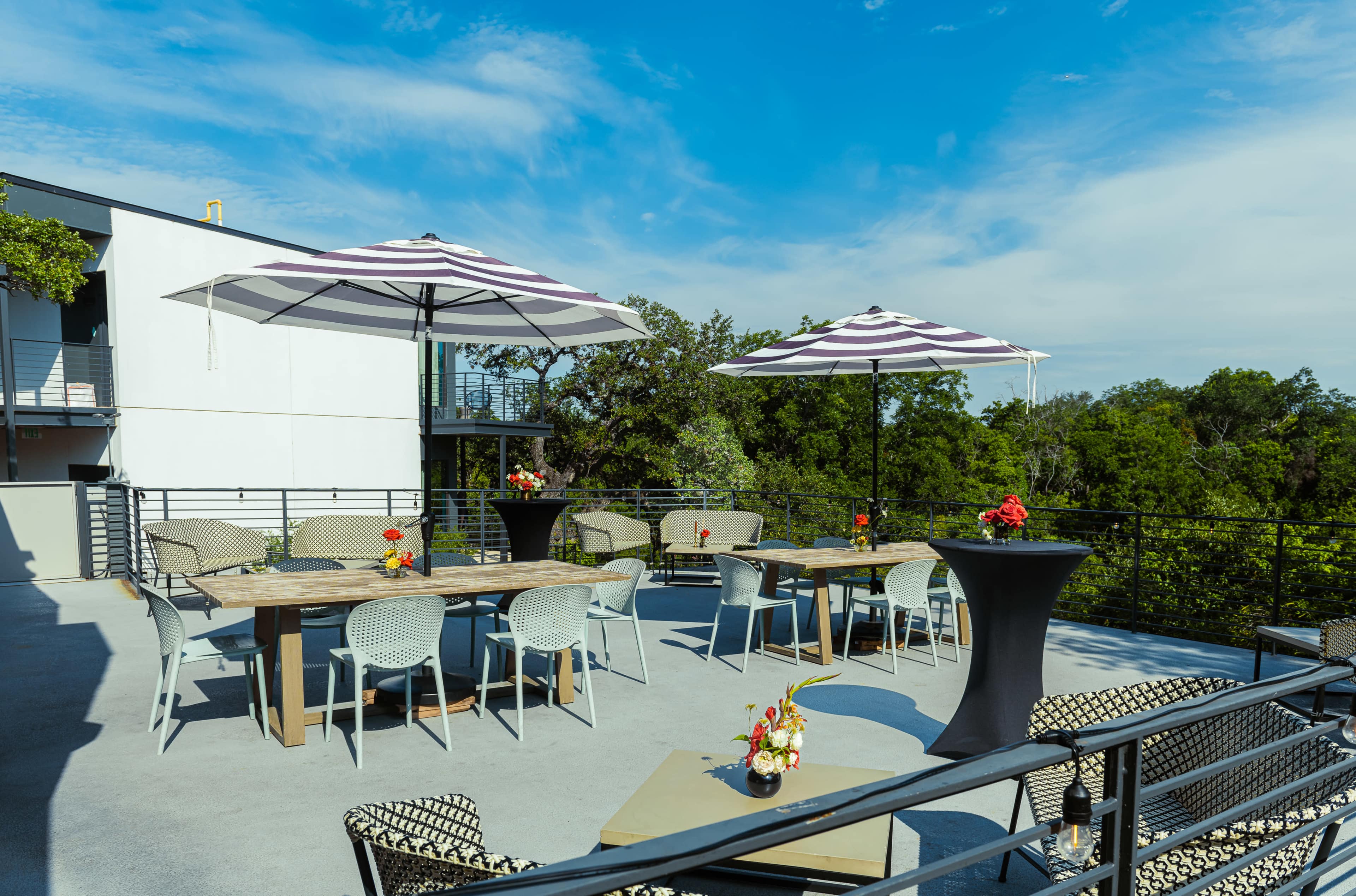 A rooftop terrace features tables with umbrellas, chairs, and decorative flower arrangements surrounded by greenery.