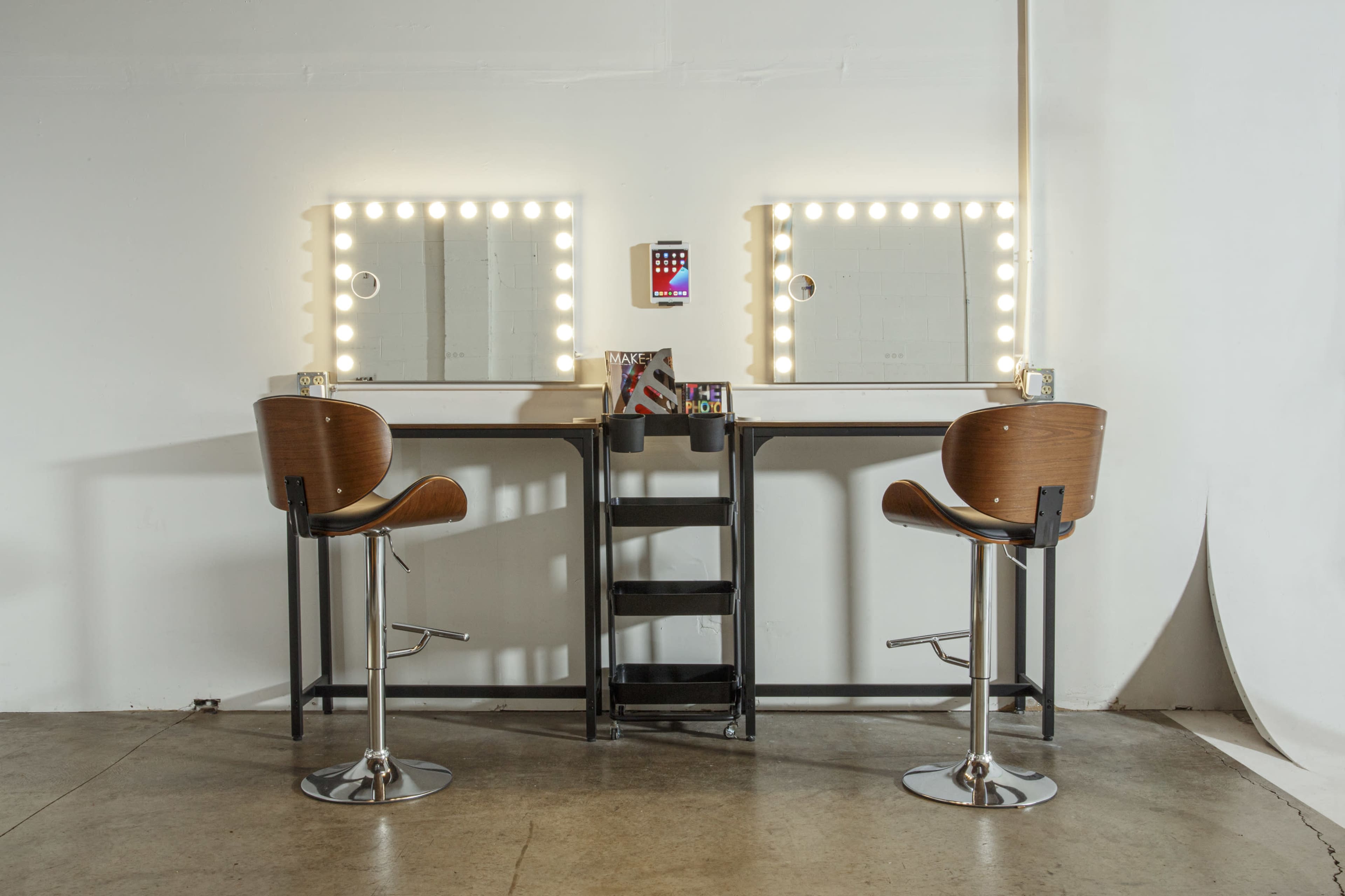 Two modern vanity tables with illuminated mirrors and high-backed stools are positioned opposite each other in a minimalist setting.