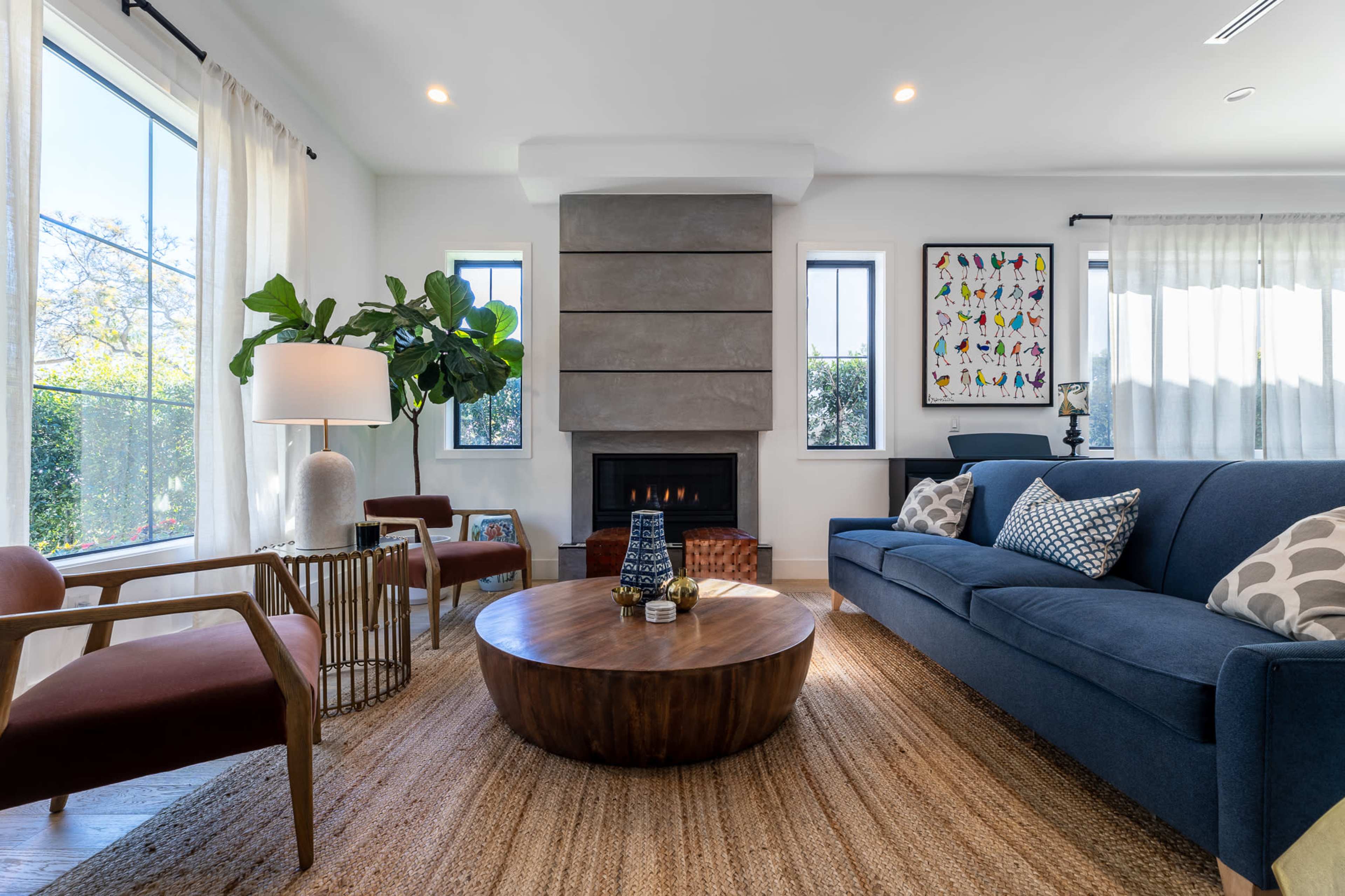 The living room features a blue sofa, a round wooden coffee table, a modern fireplace, and large windows with light curtains.