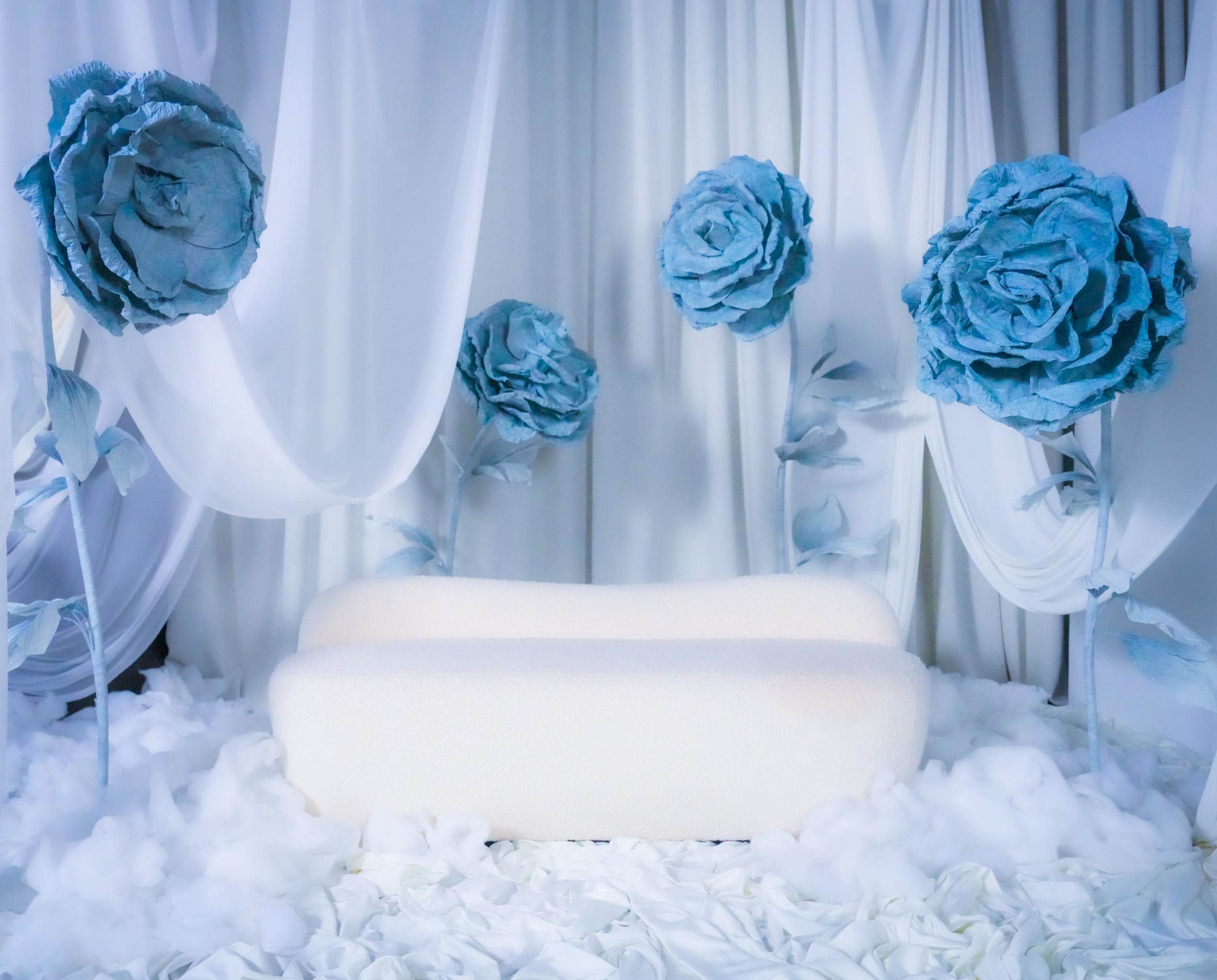 The image shows a minimalist setting featuring a white couch surrounded by large blue paper flowers on tall stems, with draped fabrics and soft white materials on the ground.