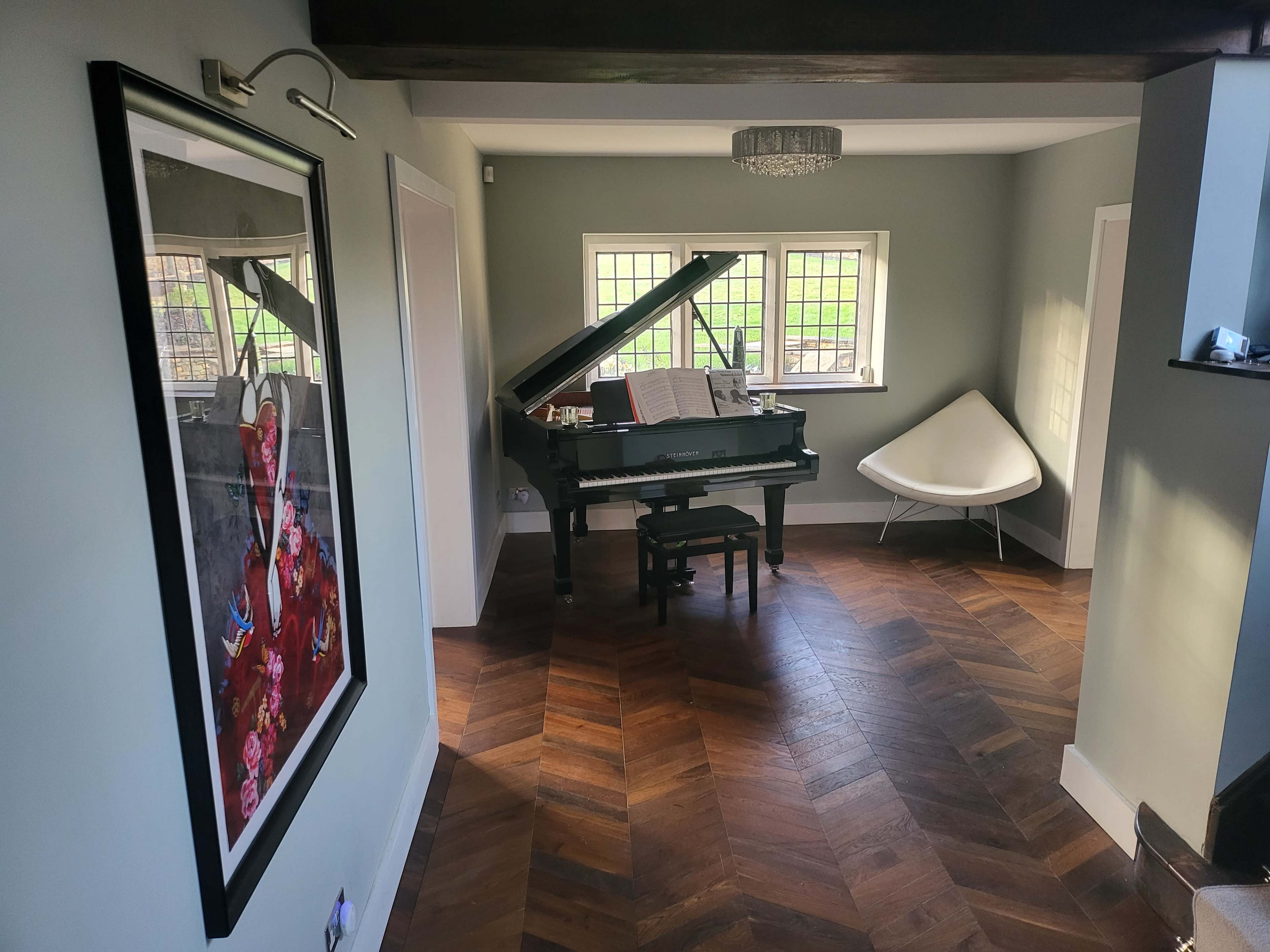 A grand piano is positioned in a well-lit room with a decorative painting on the wall and a modern chair in the corner.