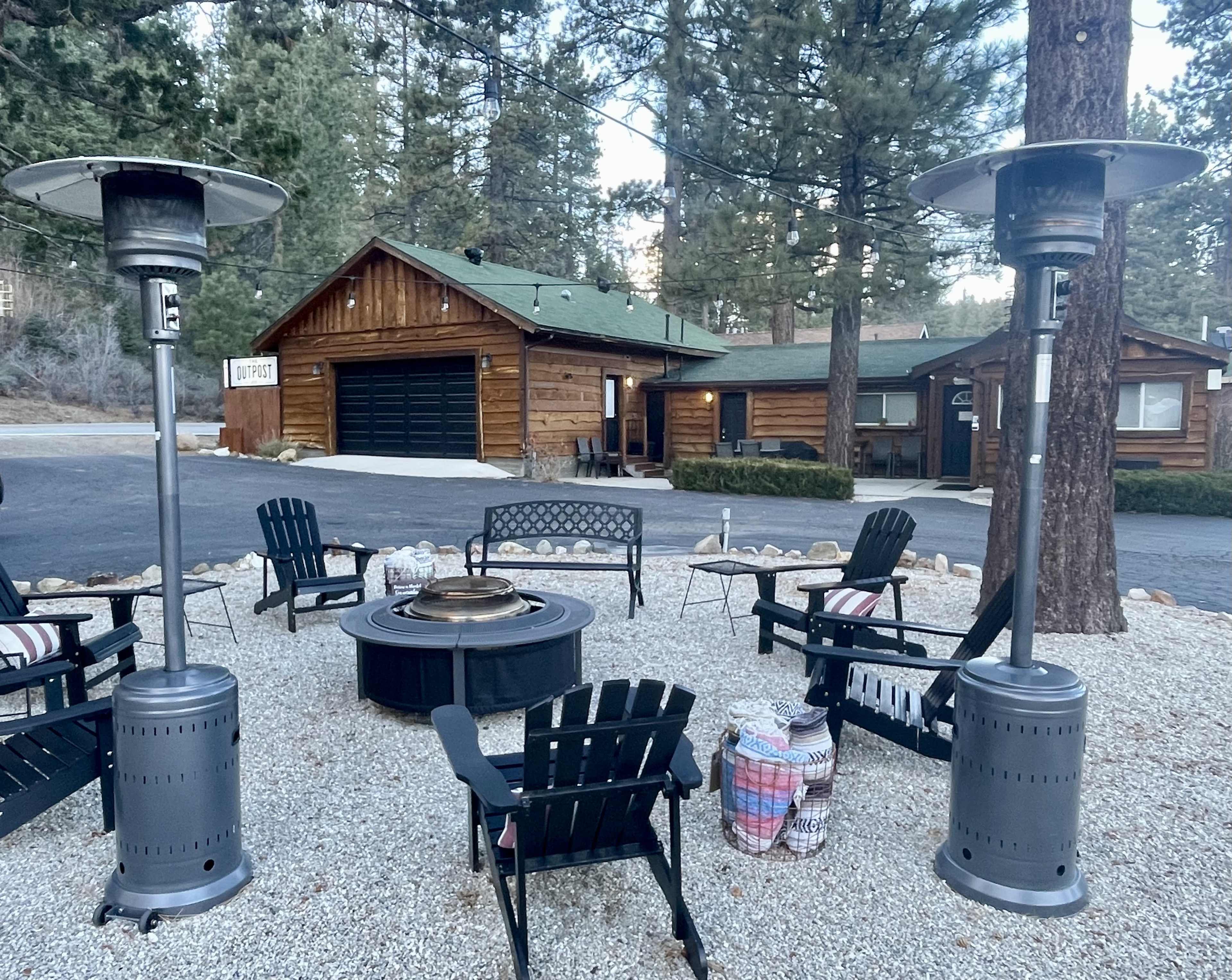 The scene features a cozy outdoor seating area with two patio heaters, a fire pit, and wooden chairs surrounded by gravel, near a log cabin structure in a forested setting.