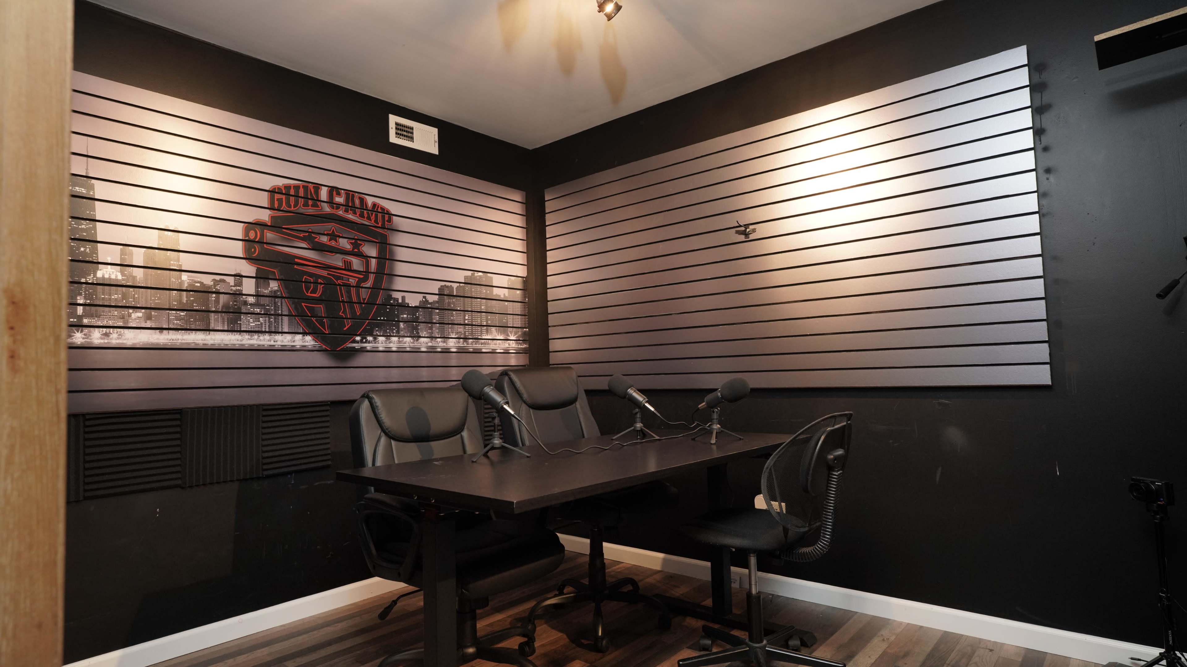 A podcast or recording studio with a dark wall, a table set with three chairs, and microphones arranged for recording.