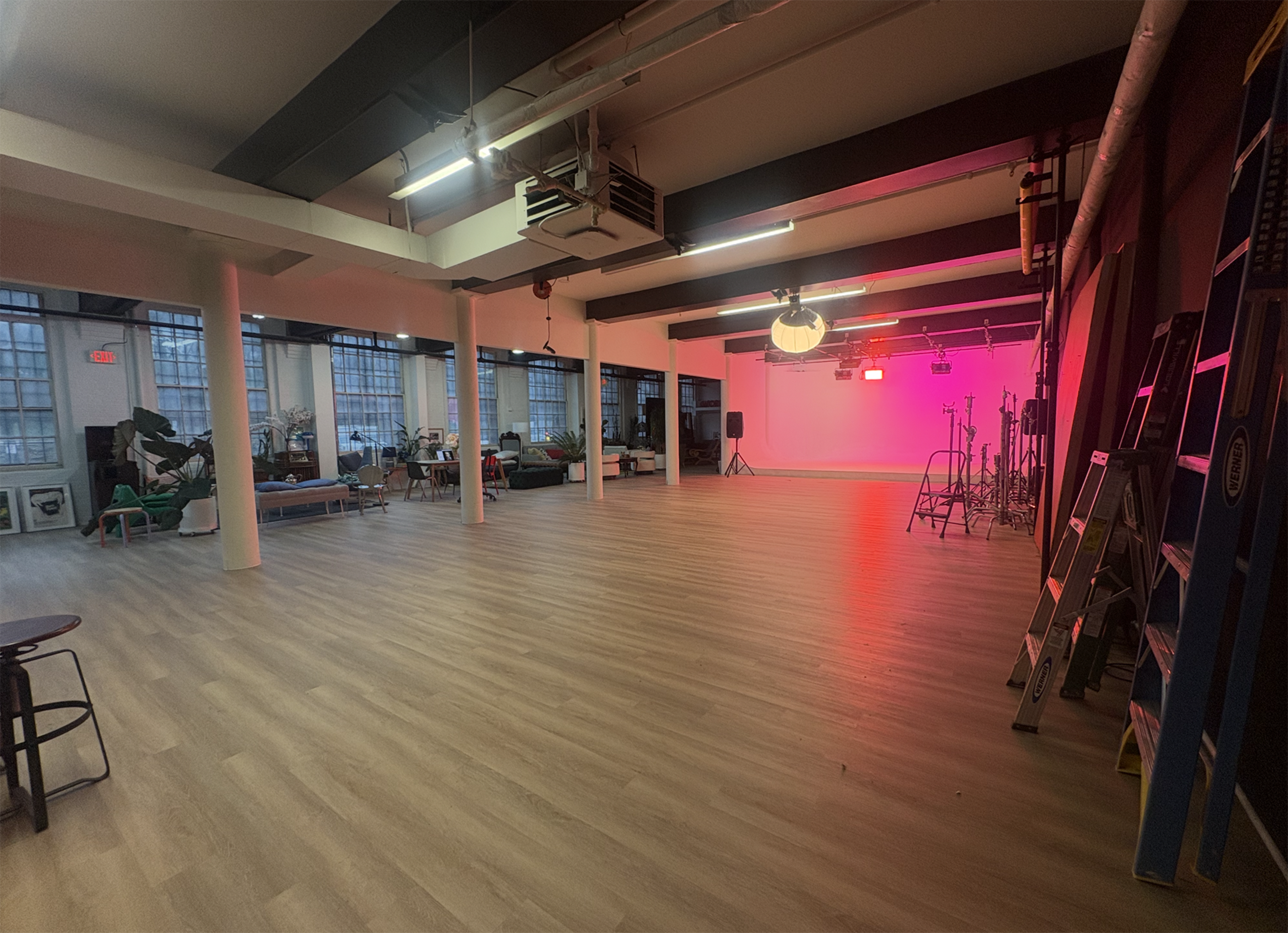 The image shows a spacious, empty studio with wooden flooring and various equipment arranged along the walls, illuminated by pink lighting.