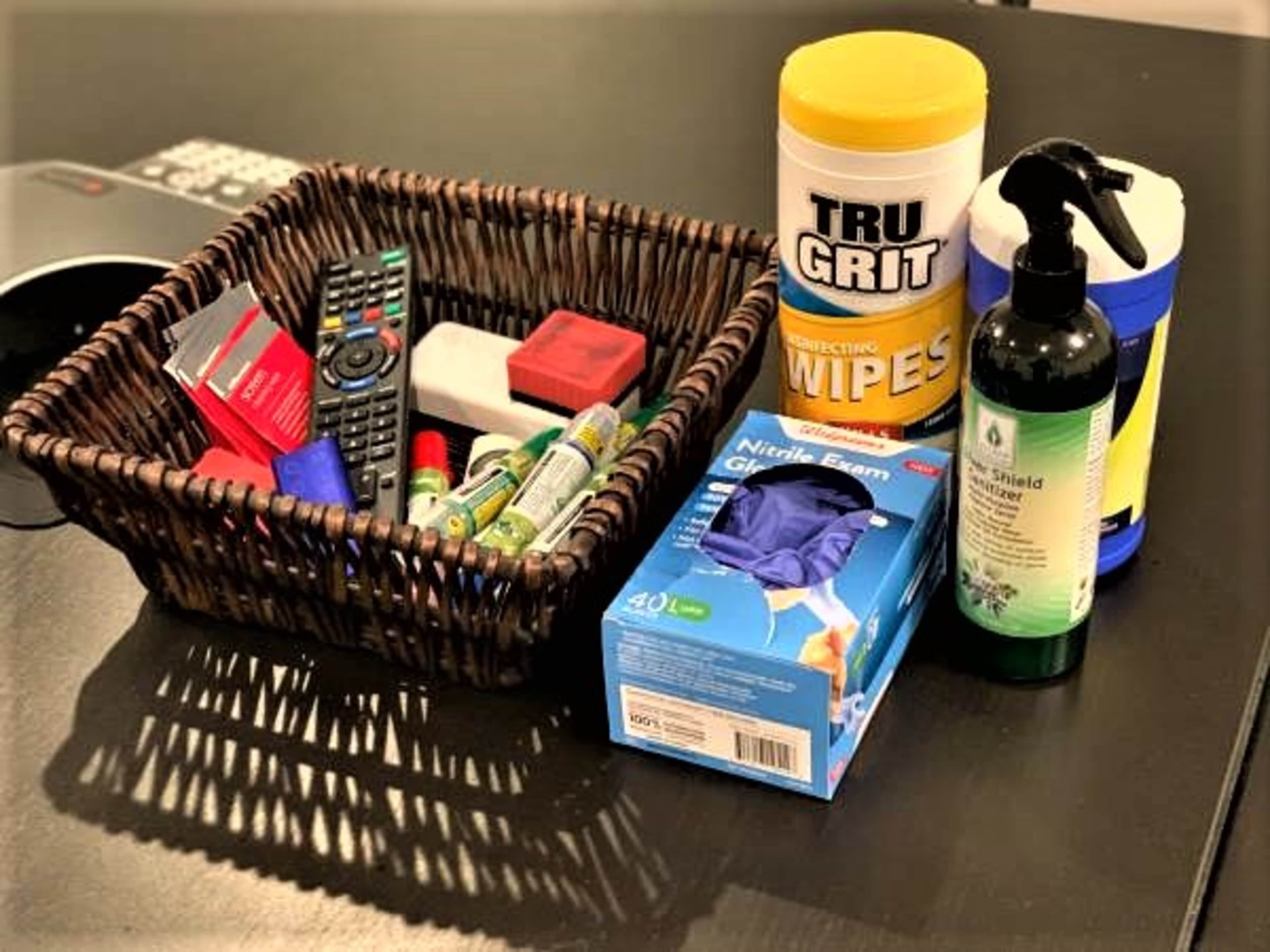 A wicker basket containing various items, including a remote control, cleaning wipes, gloves, and small containers, arranged on a black surface.