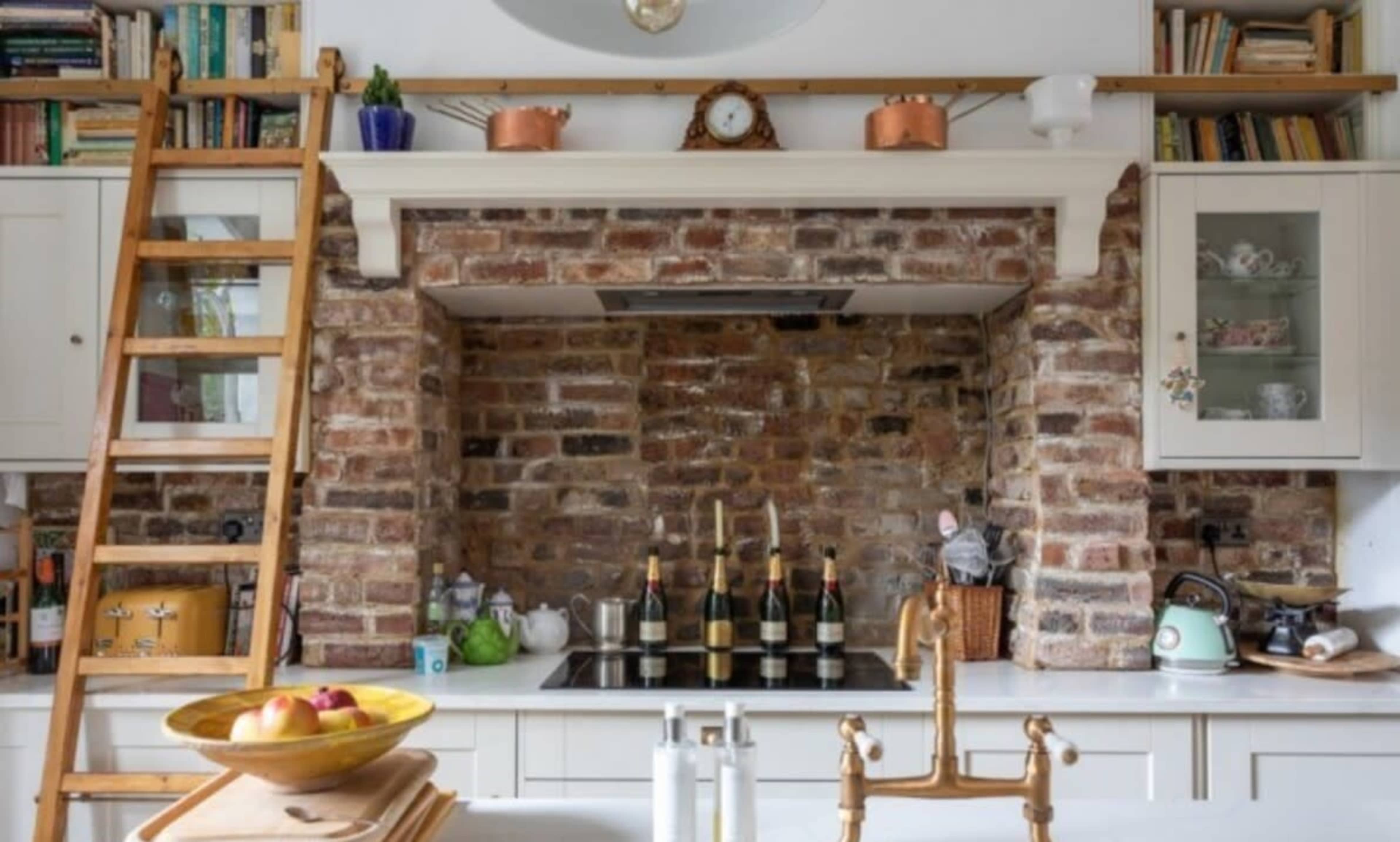 The kitchen features a rustic brick wall with a stovetop, a wooden ladder, and shelves holding various items.