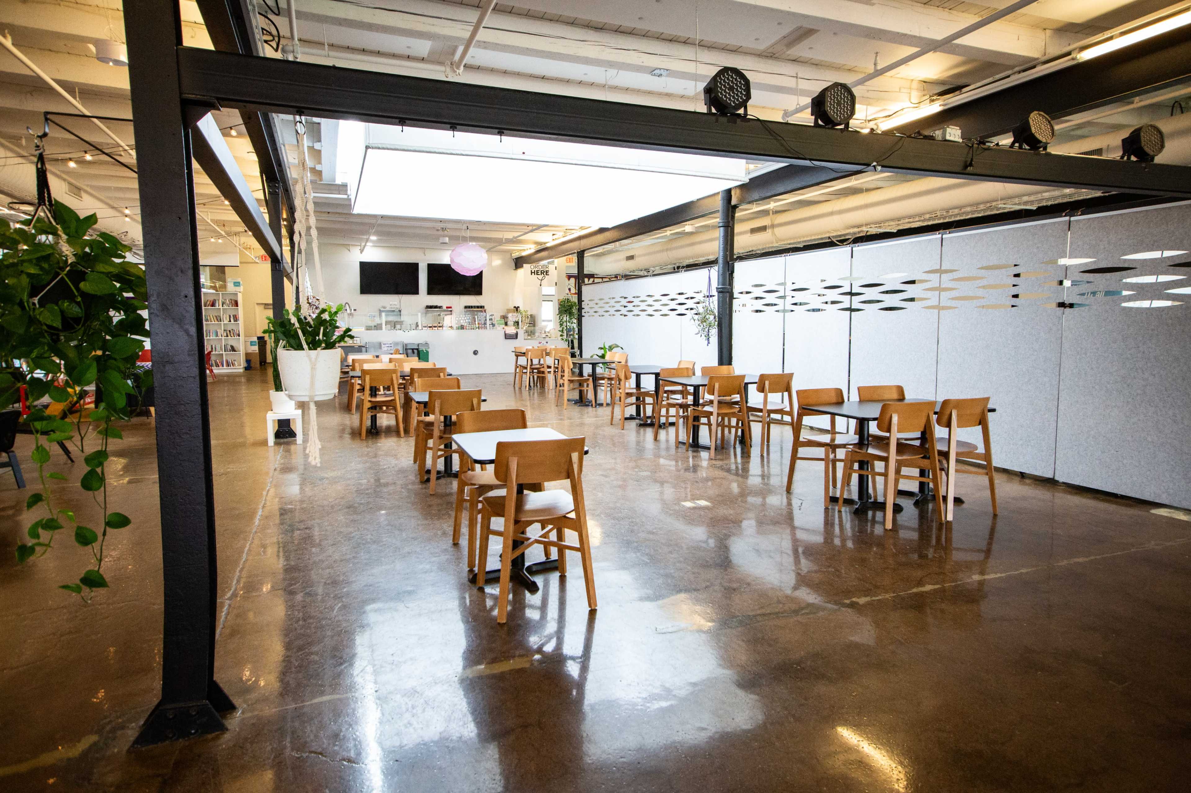 The image shows a spacious café with several wooden tables and chairs arranged in an orderly manner, alongside large windows and light-colored walls.