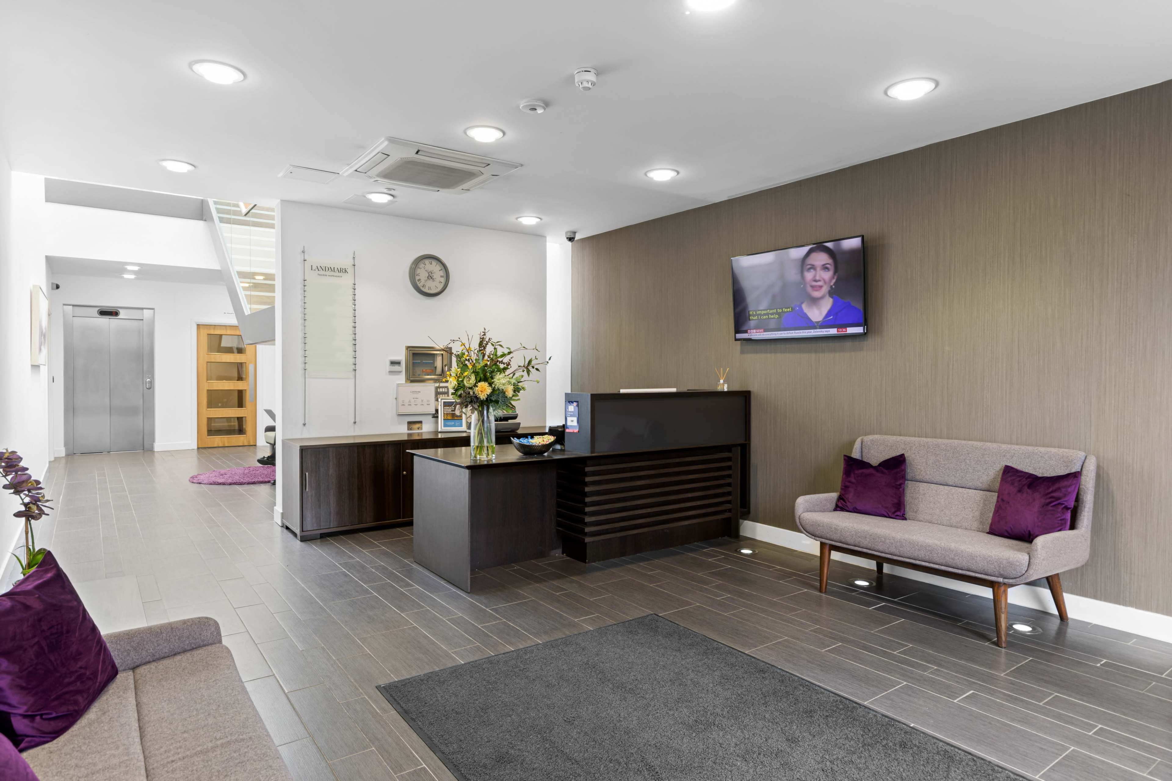 A modern reception area with a seating arrangement, a front desk, and a television displaying a video in a well-lit space.