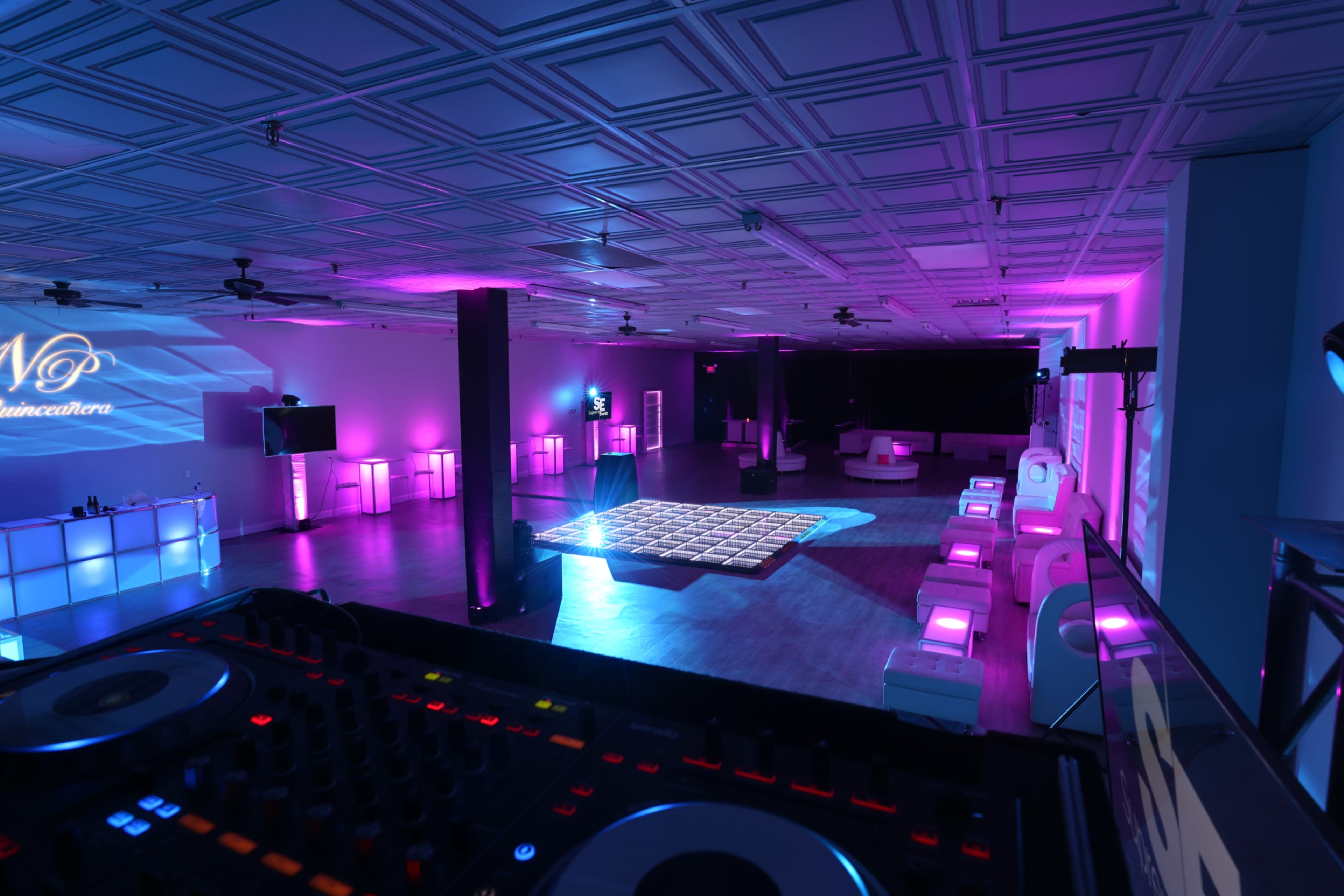 The image shows a spacious event room with a dance floor, illuminated by purple lights, featuring a DJ setup in the foreground.