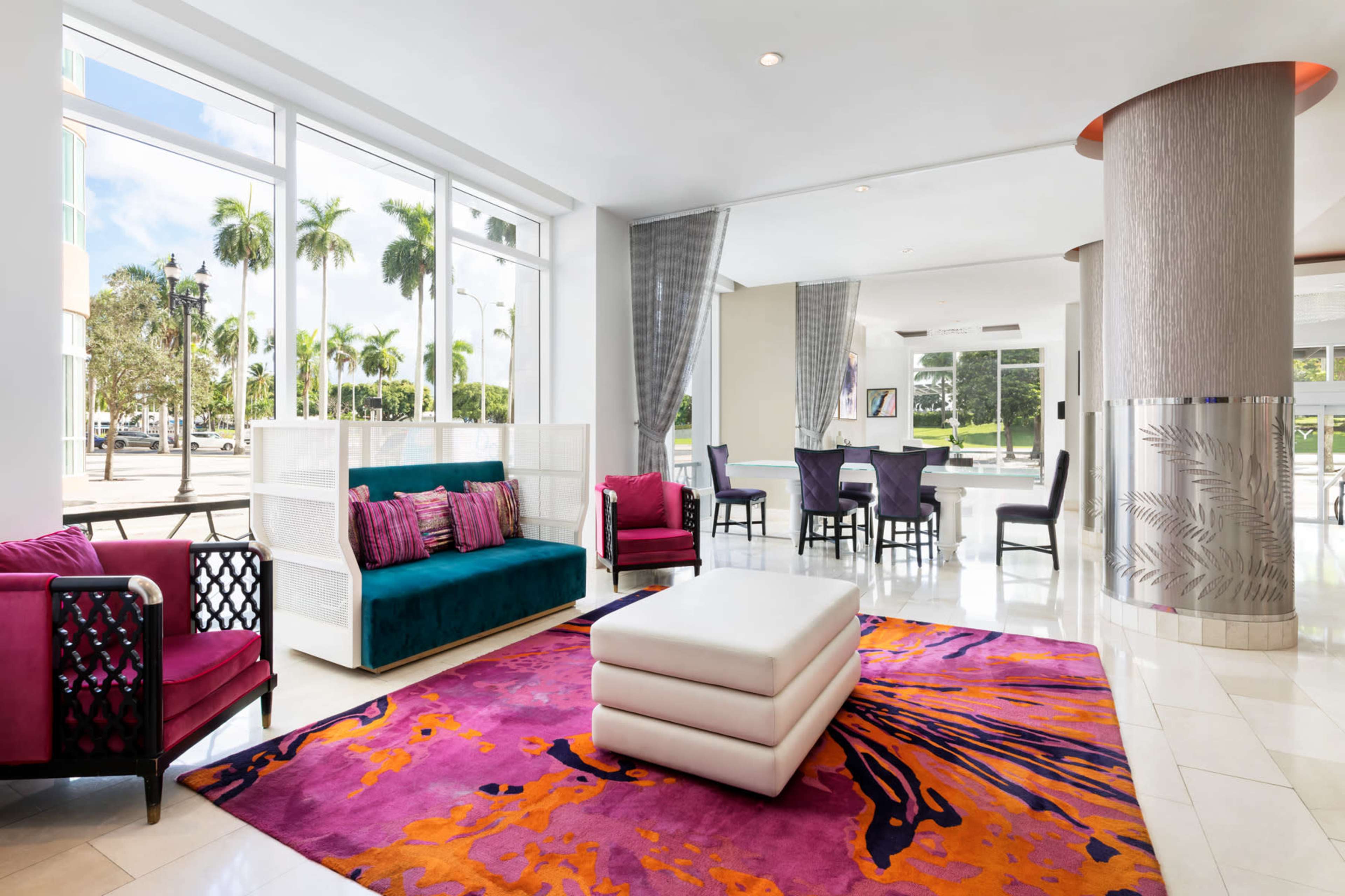 The image shows a brightly lit lobby area featuring modern seating, a large colorful rug, and palm trees visible through large windows.