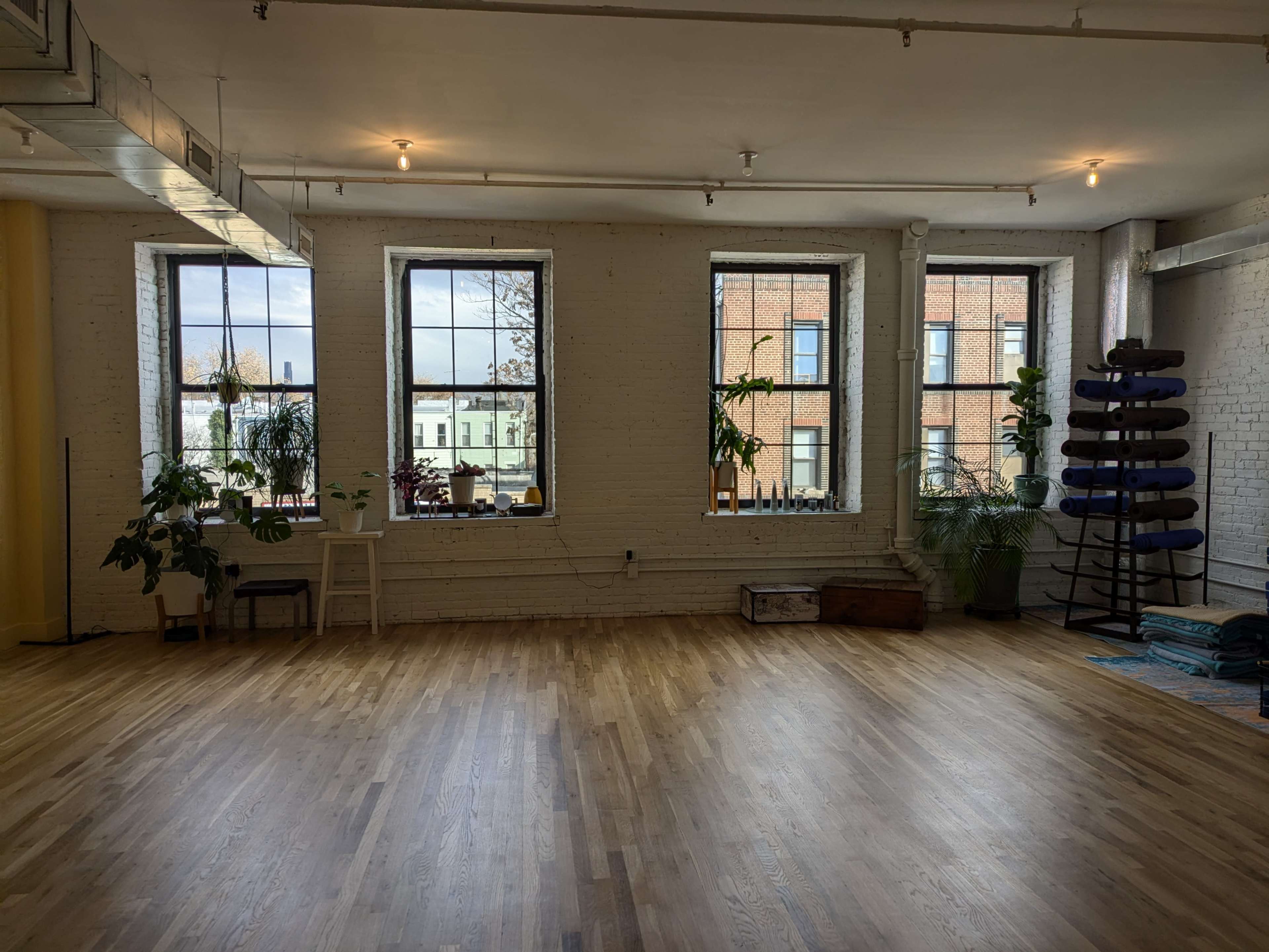 The image shows a spacious, well-lit room with large windows, plants, polished wooden floors, and yoga props arranged in a corner.