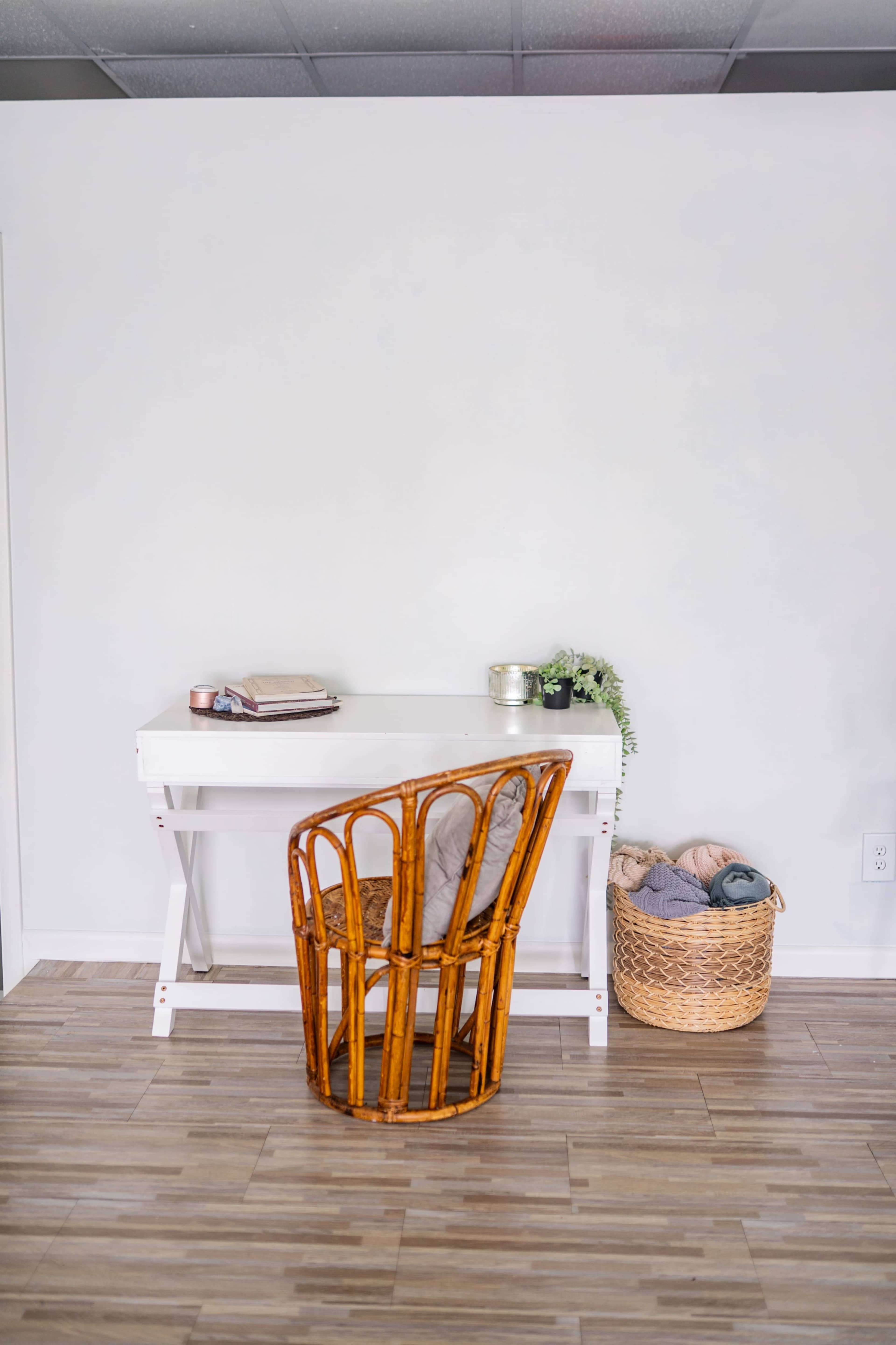 The image shows a simple white desk paired with a bamboo-style chair, positioned against a plain wall, with a woven basket containing folded textiles nearby.