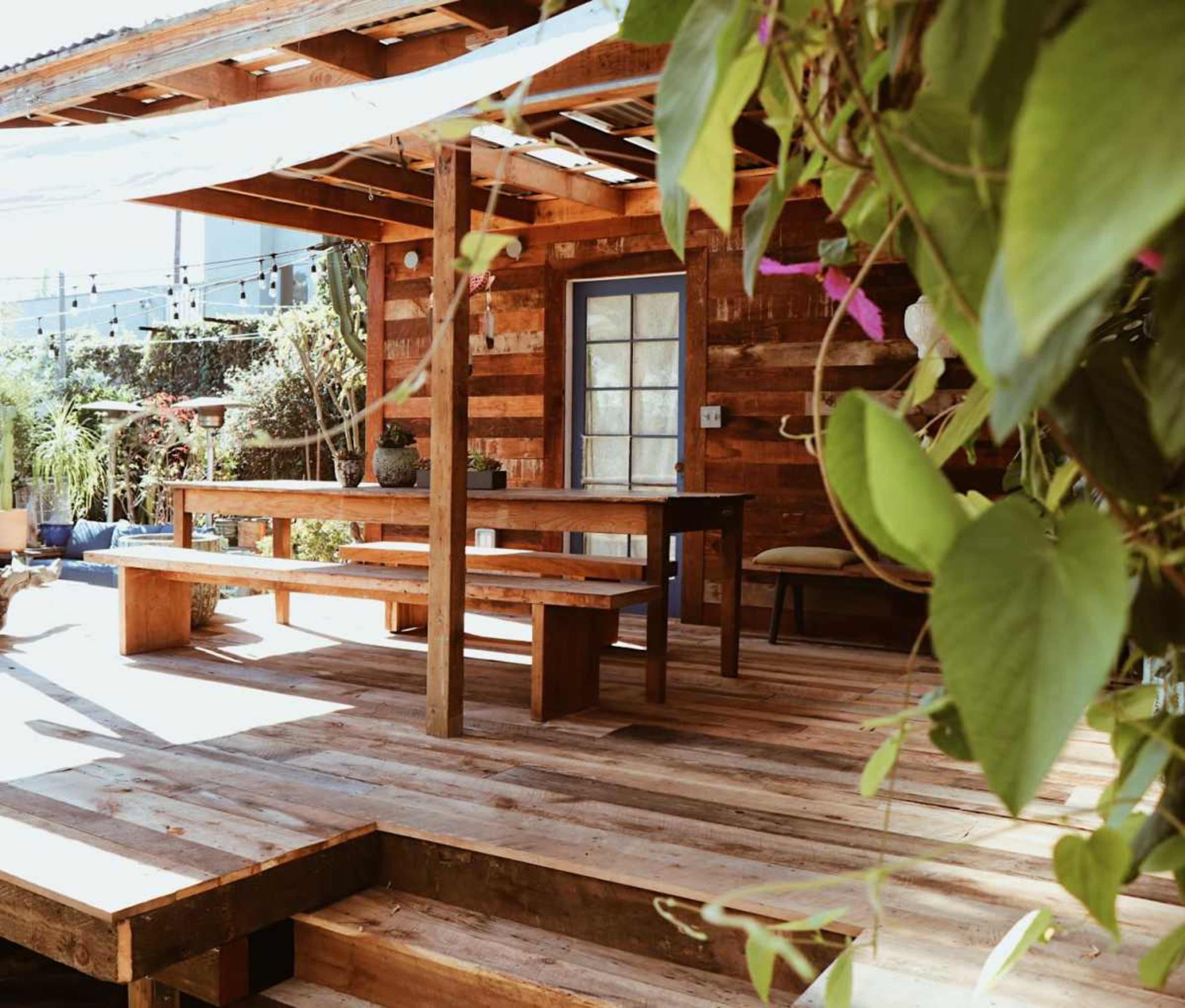 The image shows a wooden patio with a table and benches, surrounded by plants and natural light.