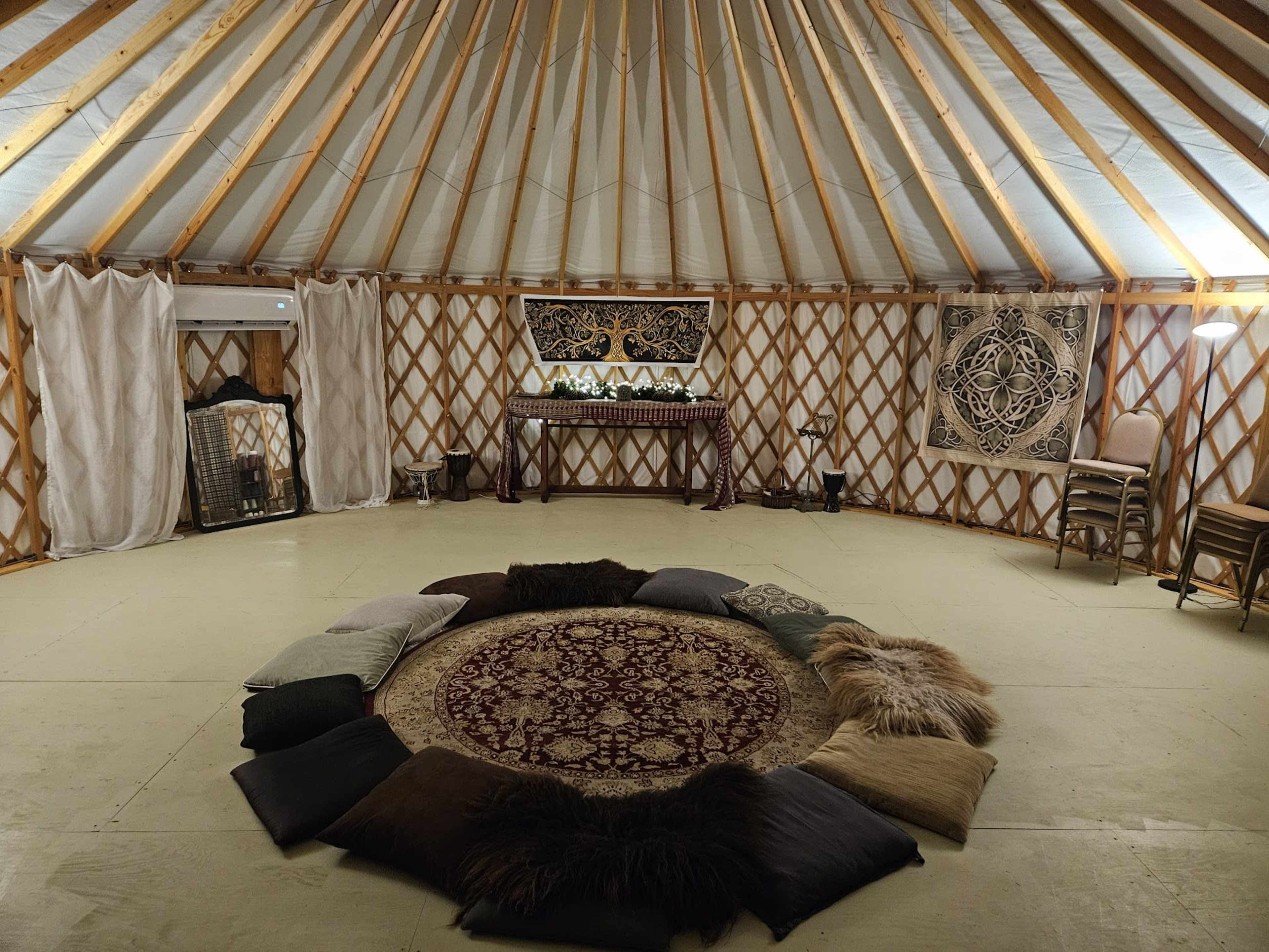 The interior of a yurt features a circular arrangement of cushions around a decorative rug, with wooden beams supporting the ceiling and various furnishings along the walls.