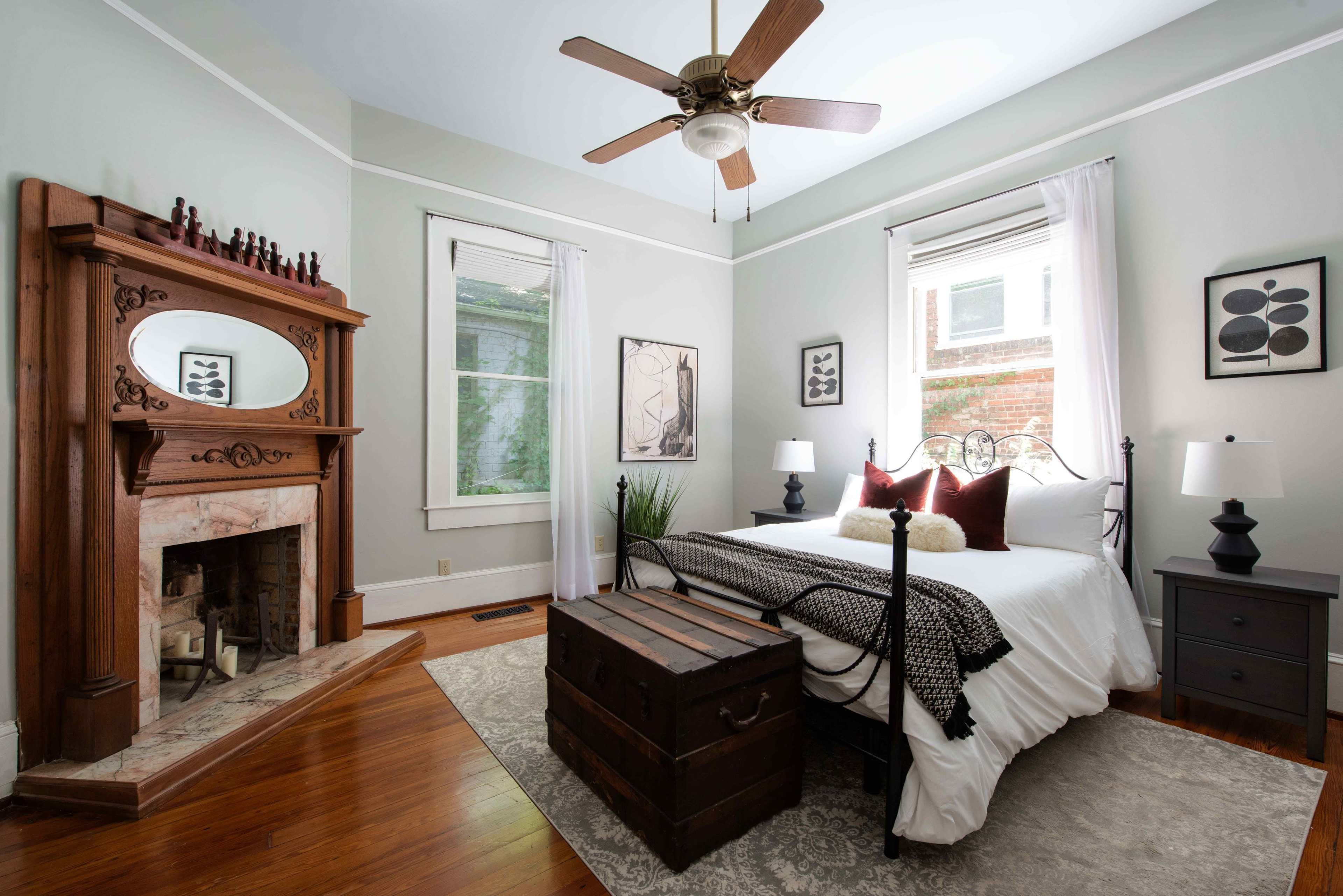 A bedroom features a bed with white linens, a wooden fireplace mantel, and a trunk at the foot of the bed, with windows letting in natural light.
