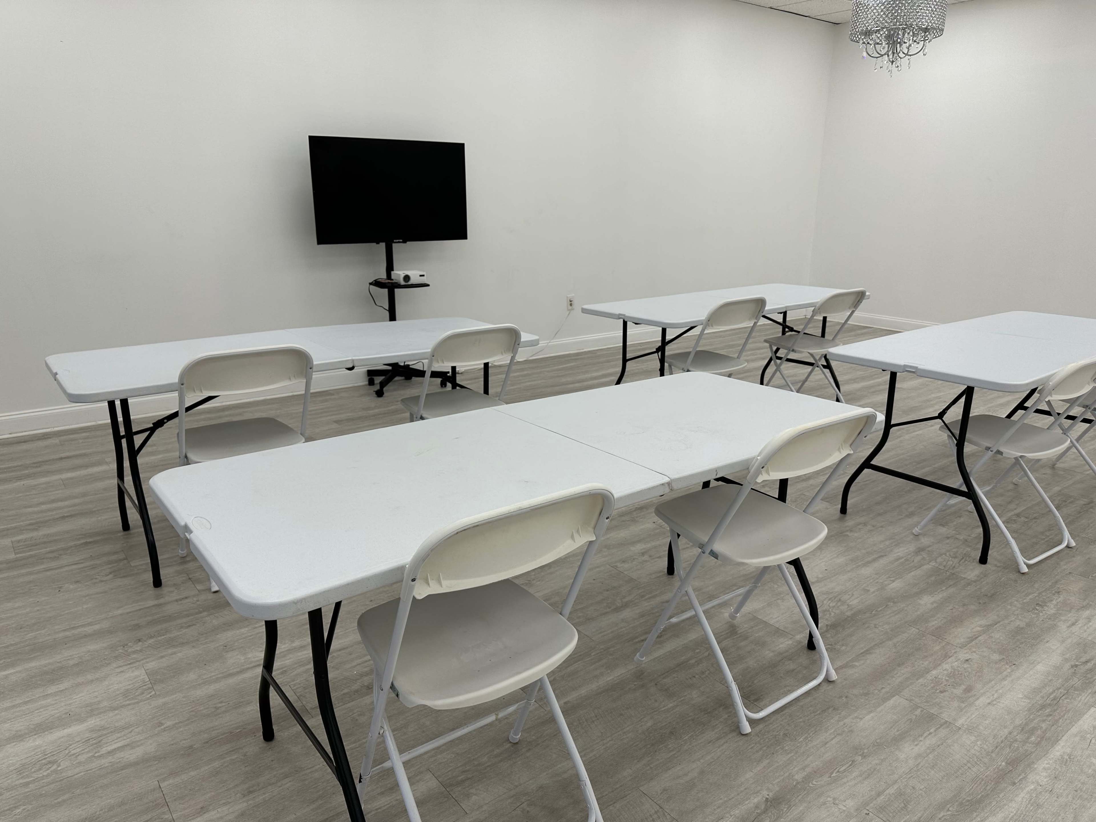 The image shows a spacious, empty meeting room with several folding tables and chairs arranged in a grid pattern, along with a television mounted on the wall.