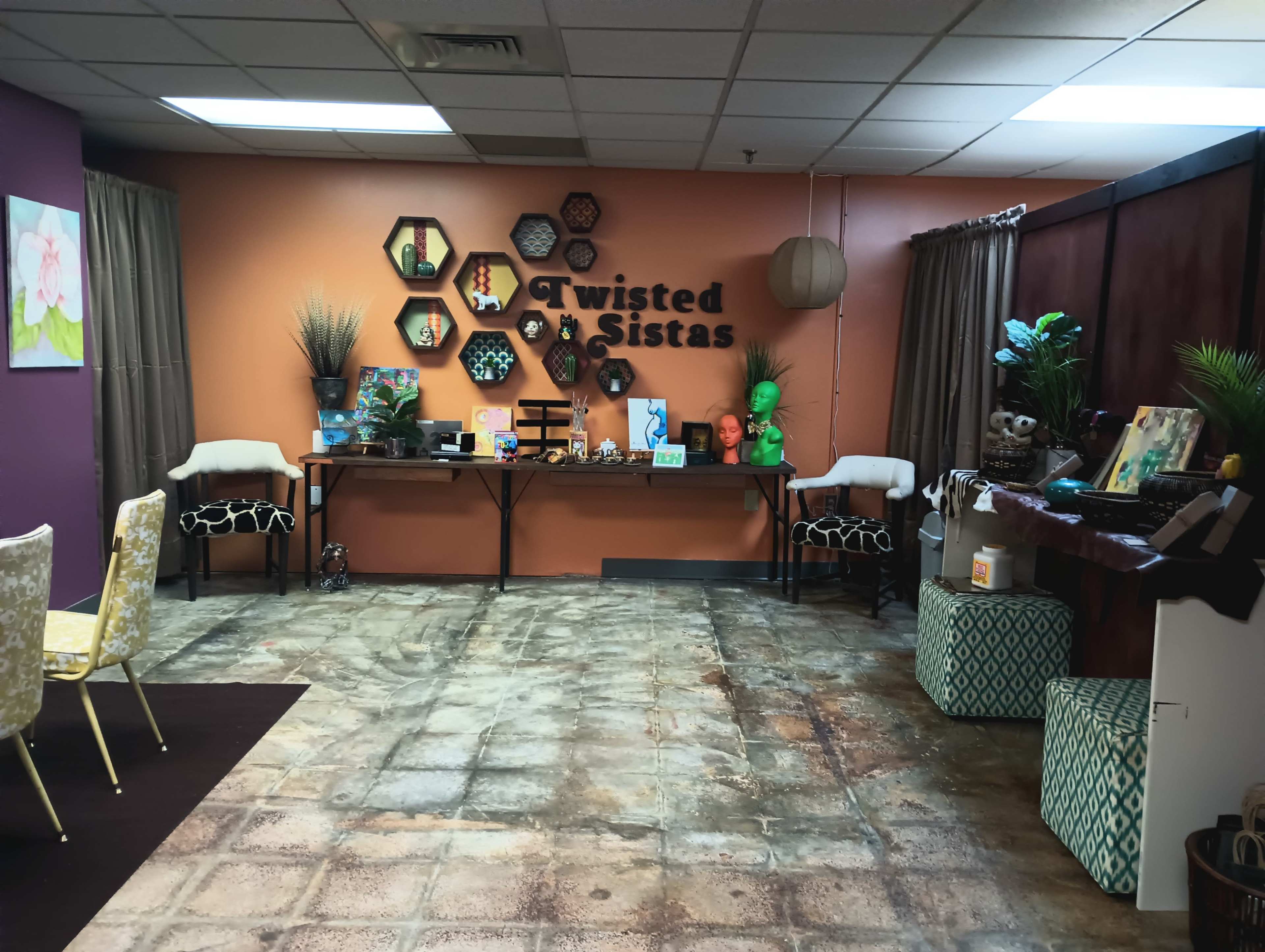 The image shows a cozy room with a decorative wall featuring the words "Twisted Sistas," along with various plants, art pieces, and a long table in the center.