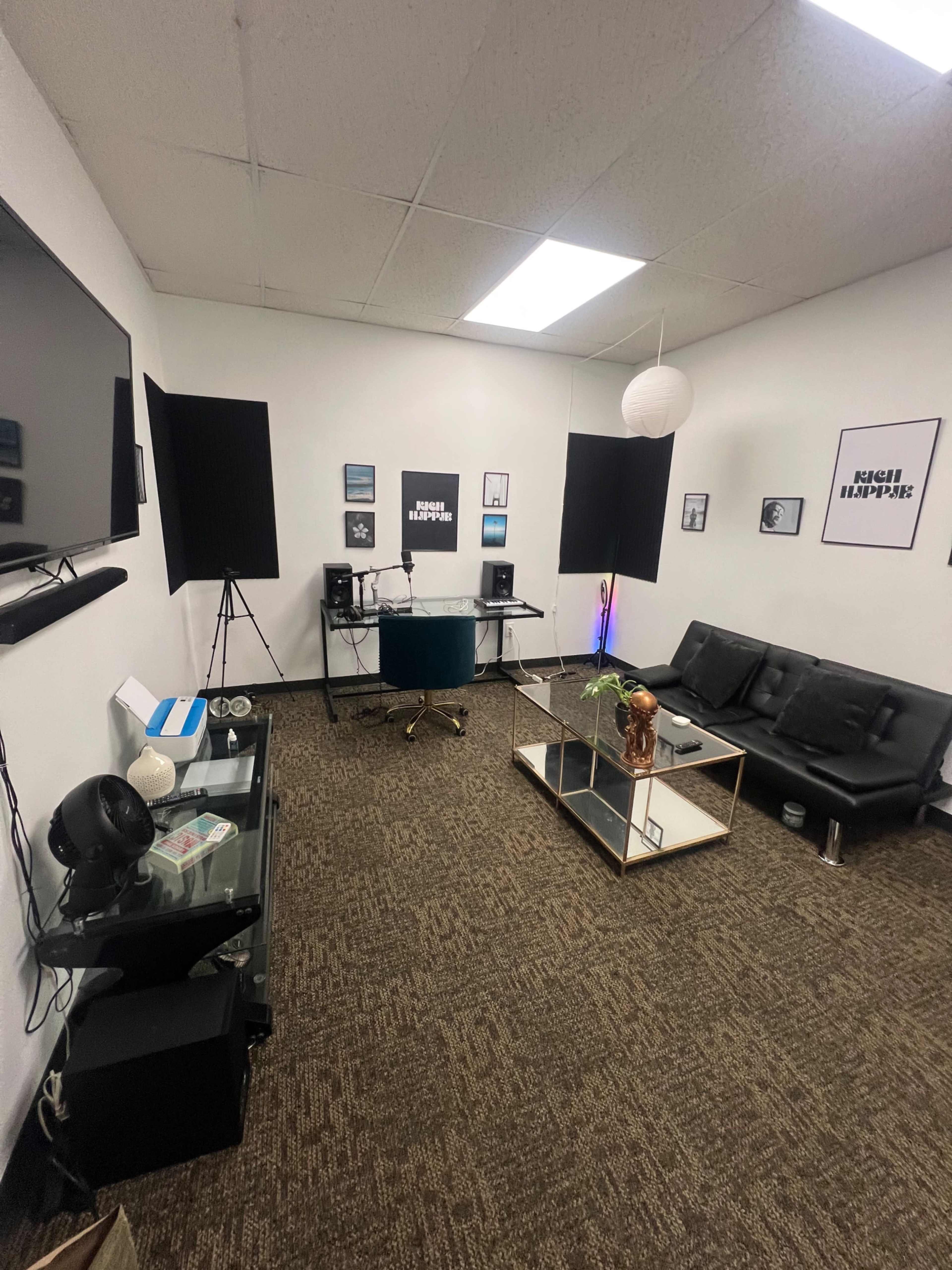 The image shows a modern recording studio or lounge area featuring a black futon, glass coffee table, desk with speakers, and various framed artworks on the walls.