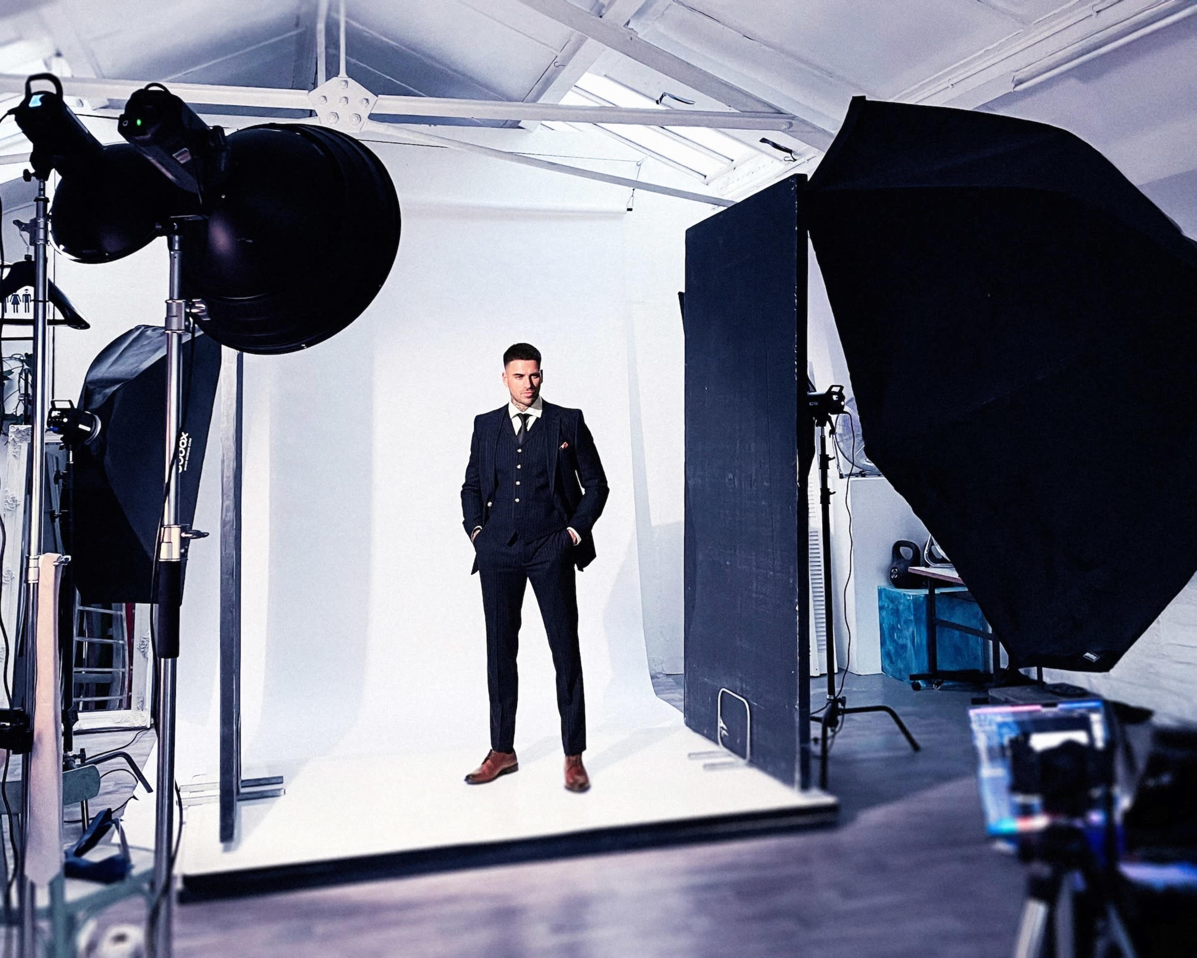 A man in a suit stands on a white backdrop inside a photography studio, surrounded by large studio lights and equipment.