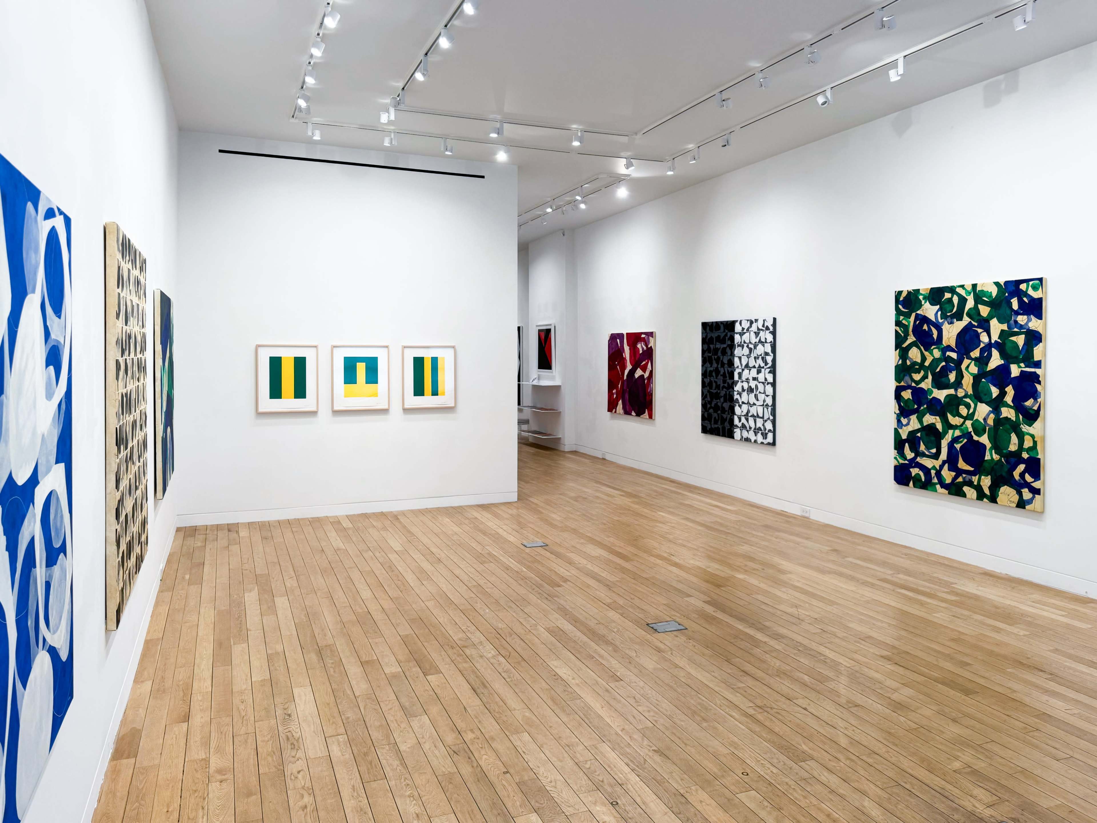 The image shows an art gallery with several colorful abstract paintings hung on white walls and a wooden floor.