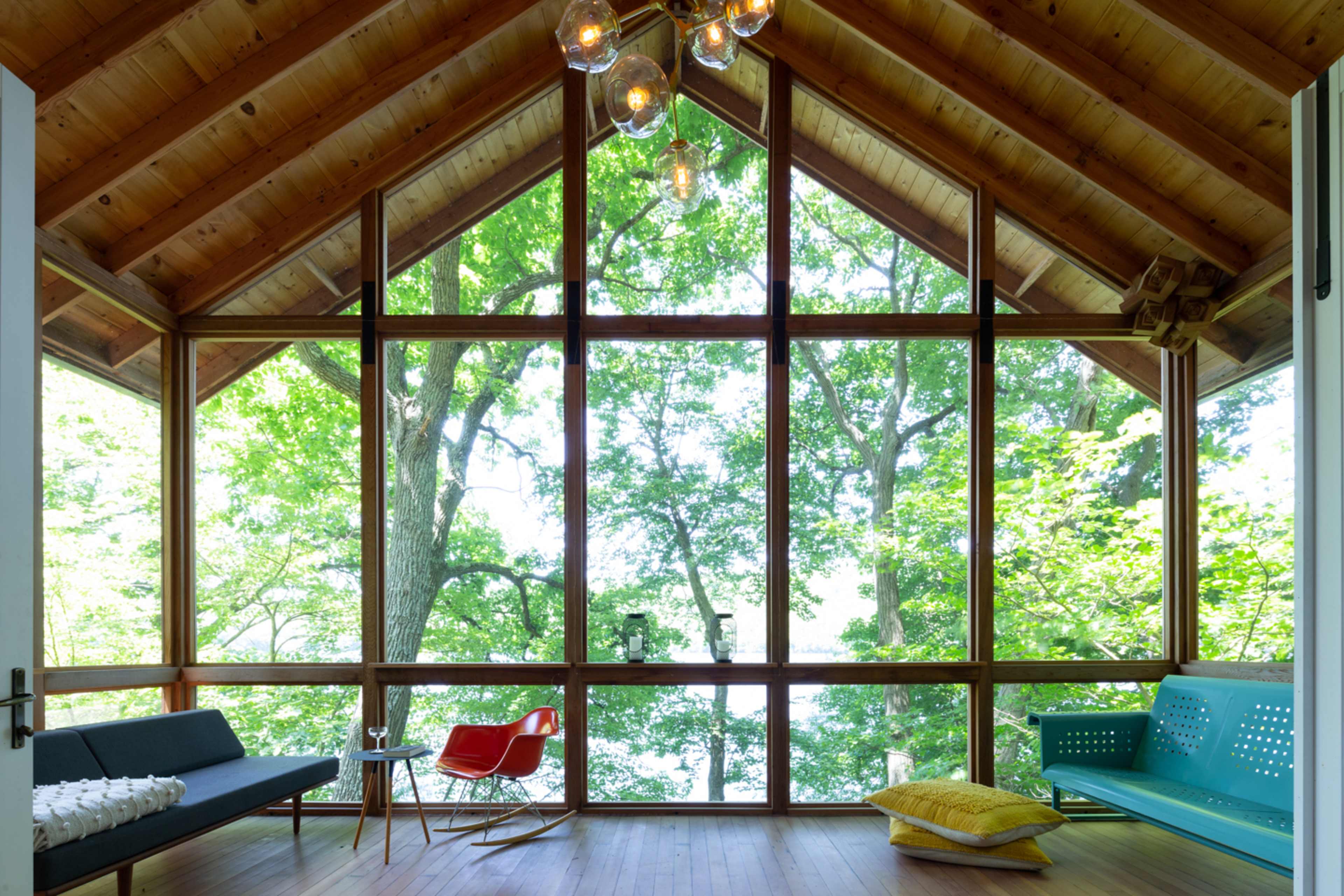 The image shows a wooden room with large windows overlooking a lush green landscape, furnished with a black couch, a red chair, and a blue bench.