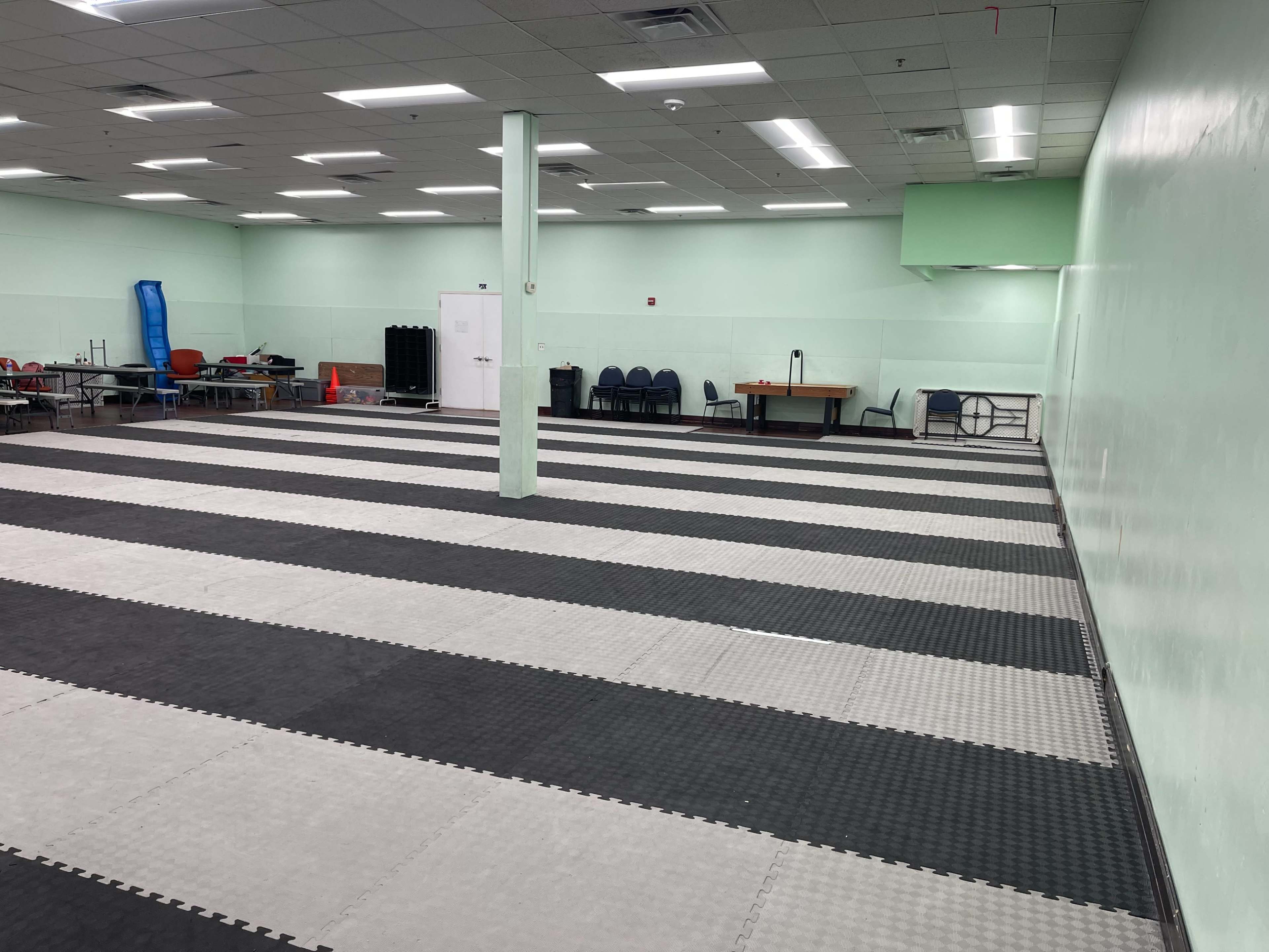 An empty training room with a checkerboard mat layout, featuring a green wall and various equipment stored at the back.