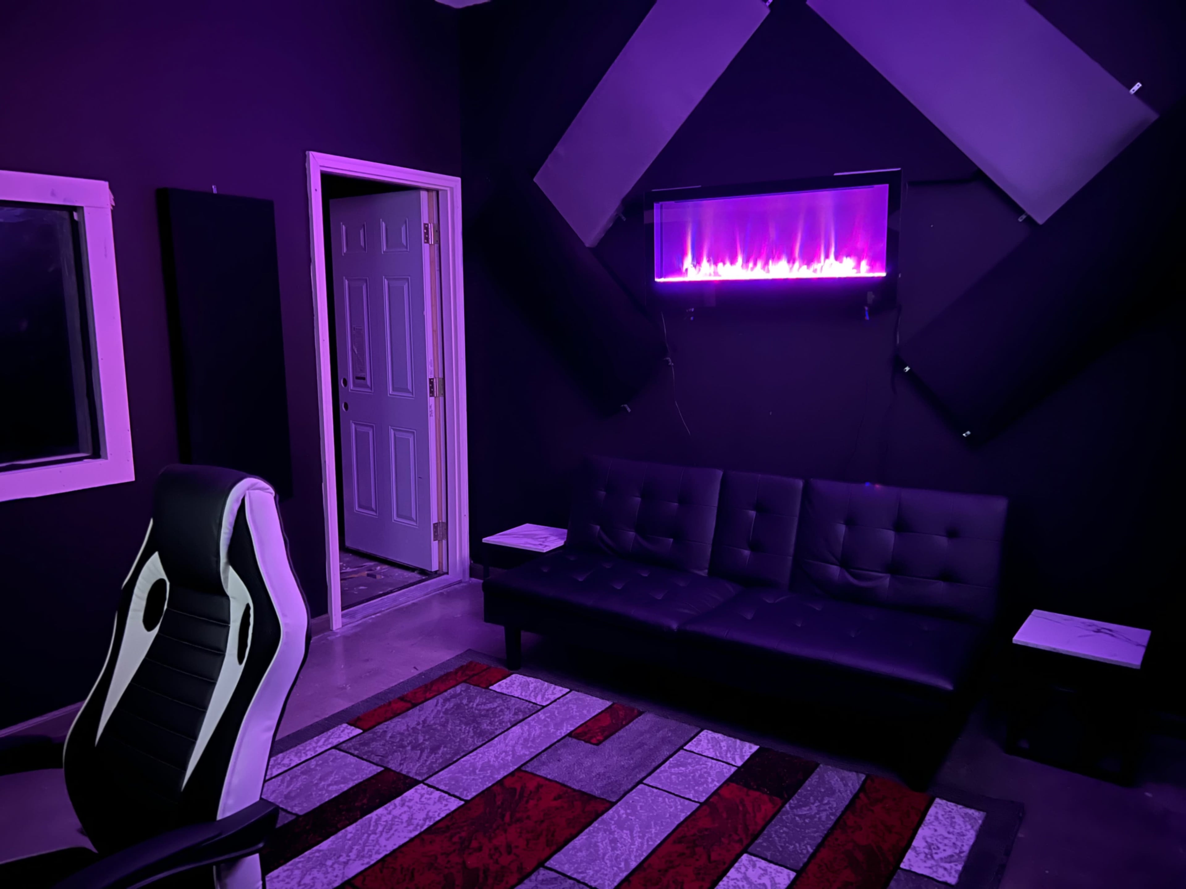A room with a black wall, a black couch, a colorful rug, and a mounted electric fireplace, featuring soundproofing panels and a door leading outside.