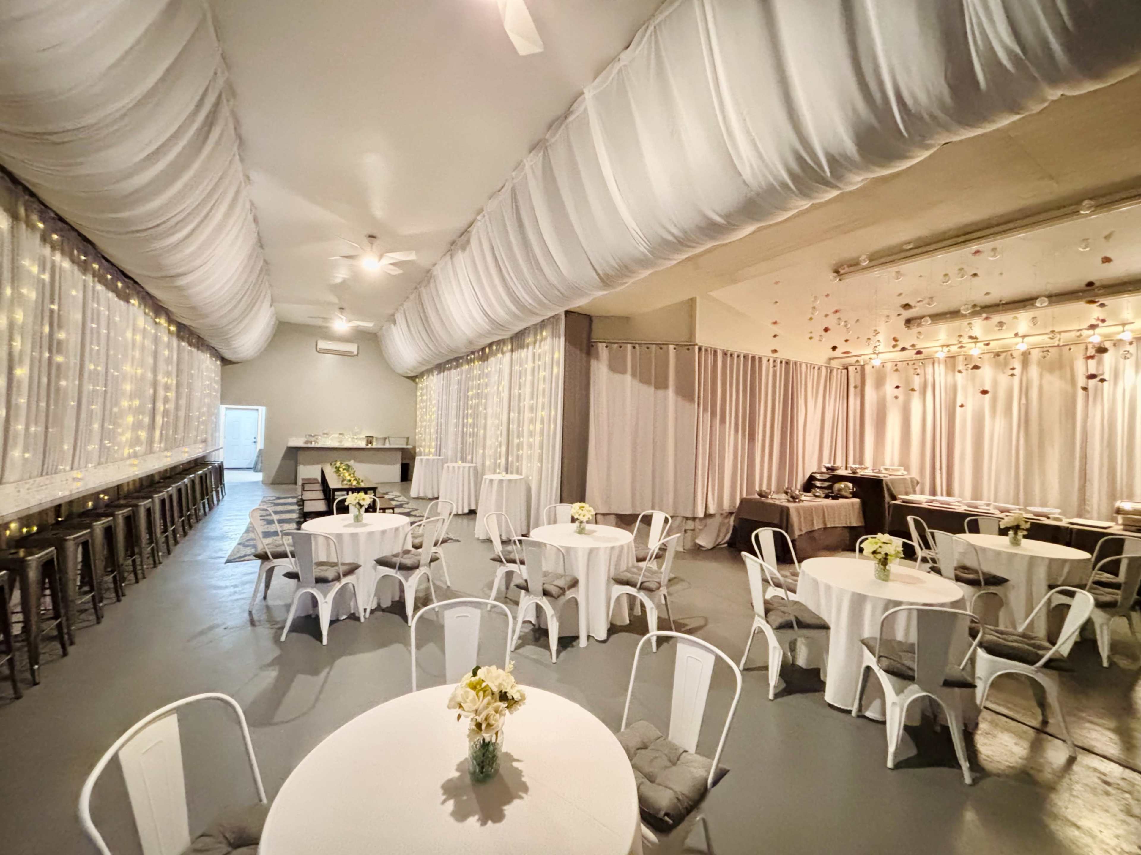 The image shows an indoor event space with tables, chairs, and draped fabric, illuminated by string lights.