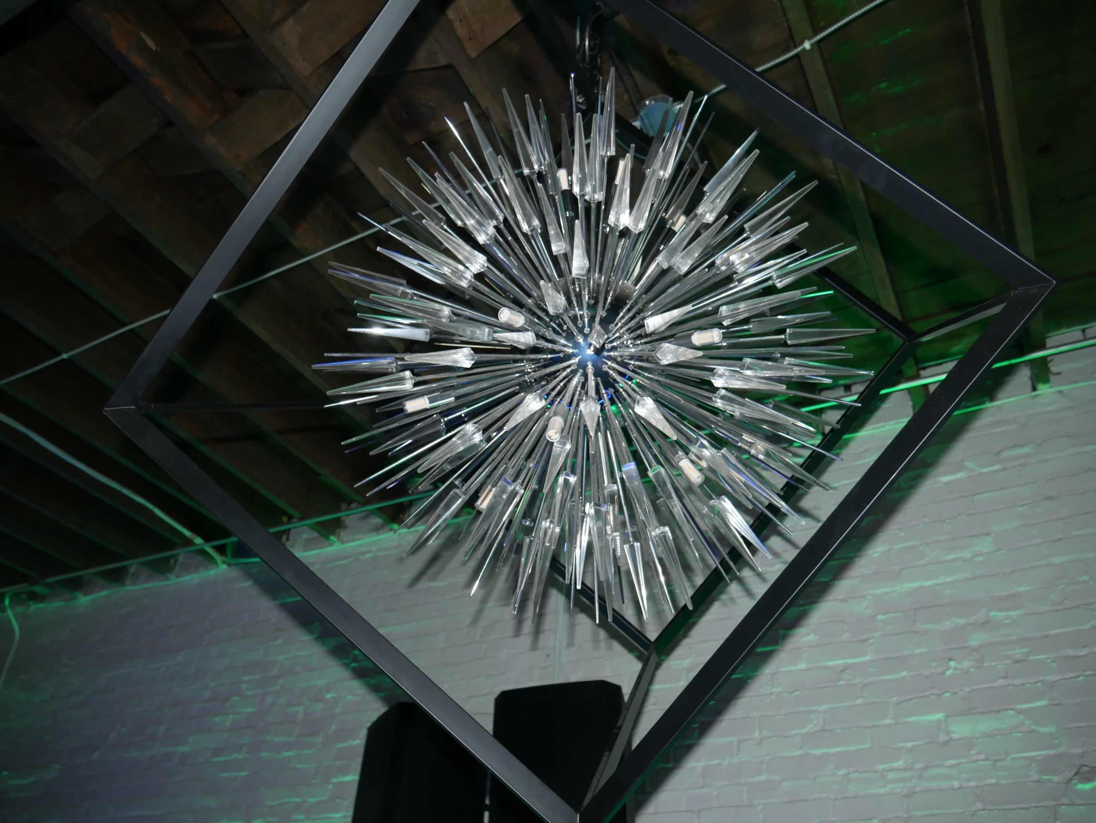 A geometric light fixture with elongated glass strands hangs from a ceiling, surrounded by a square frame.