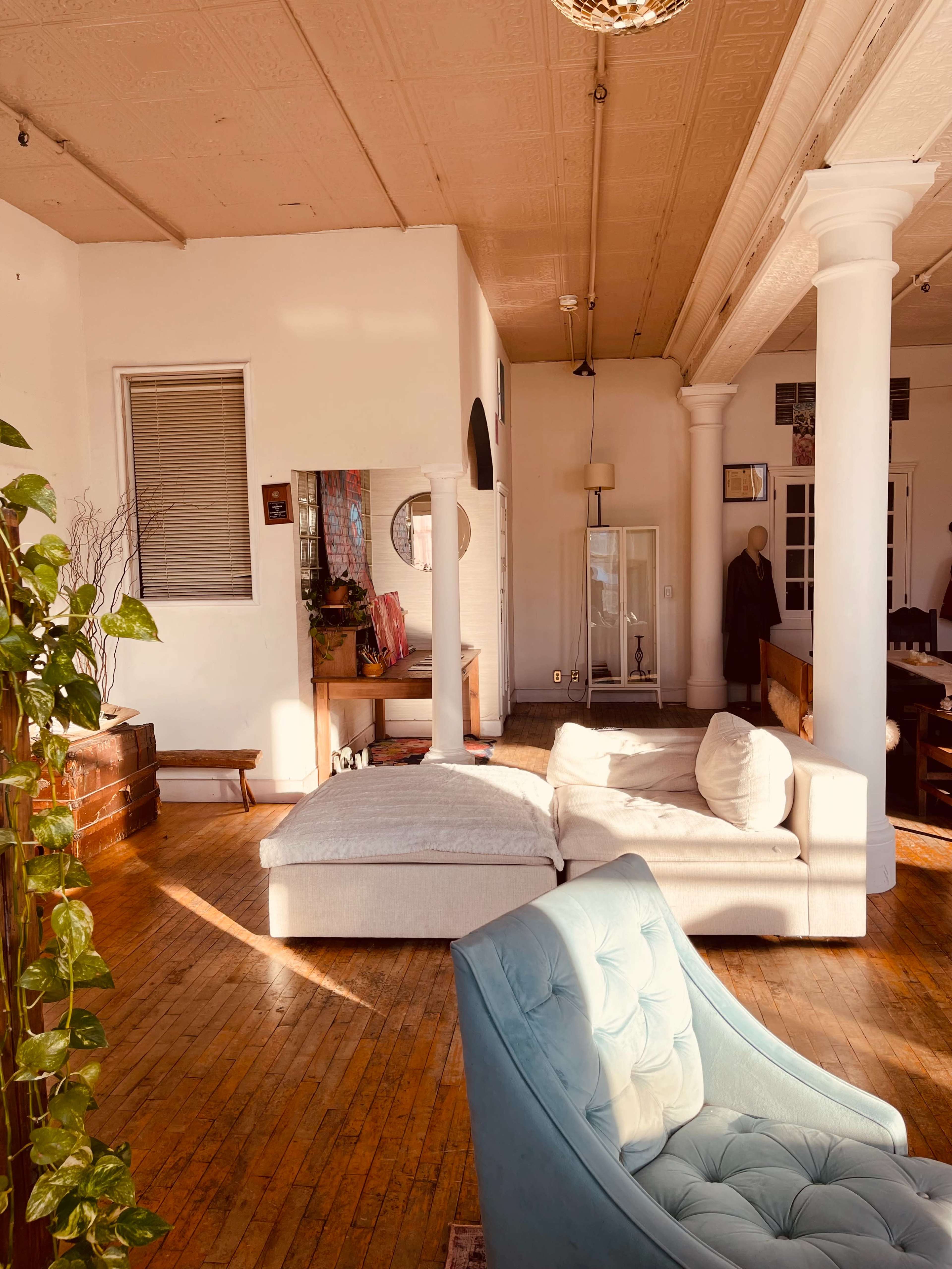 The image shows a spacious living area featuring a light-colored sectional sofa, a blue accent chair, and large pillars supporting the ceiling, illuminated by natural light.