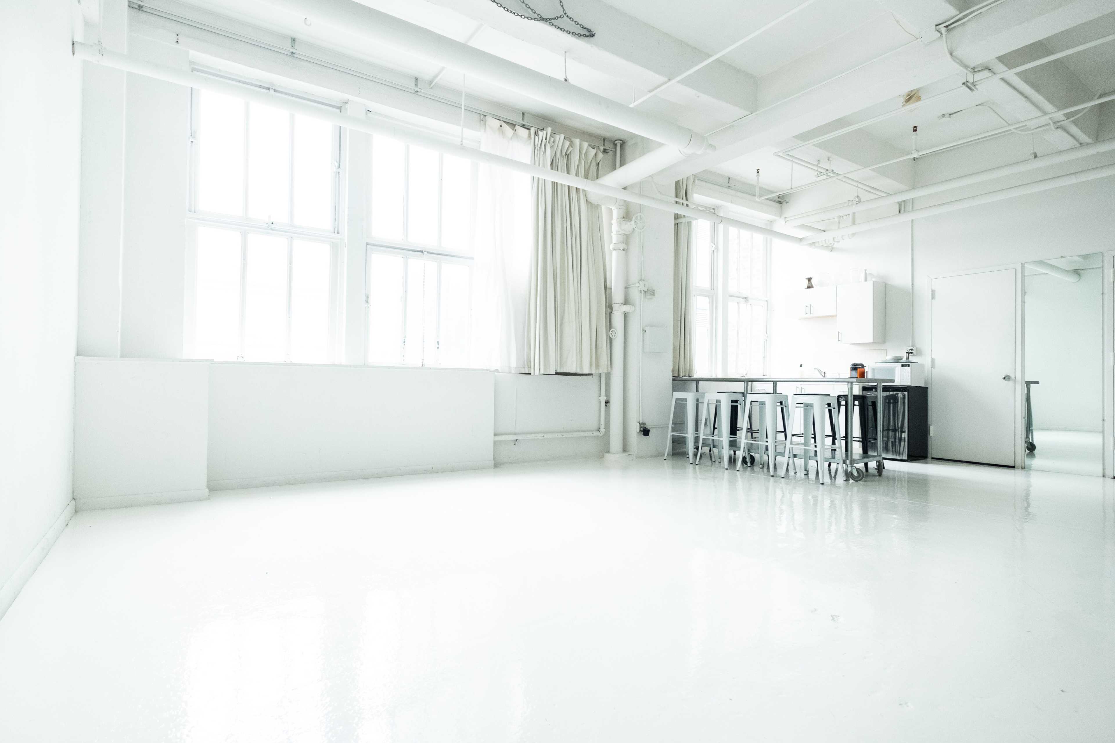 The image shows a spacious, minimalistic interior with white walls, a polished floor, large windows, and a kitchen area in the corner.