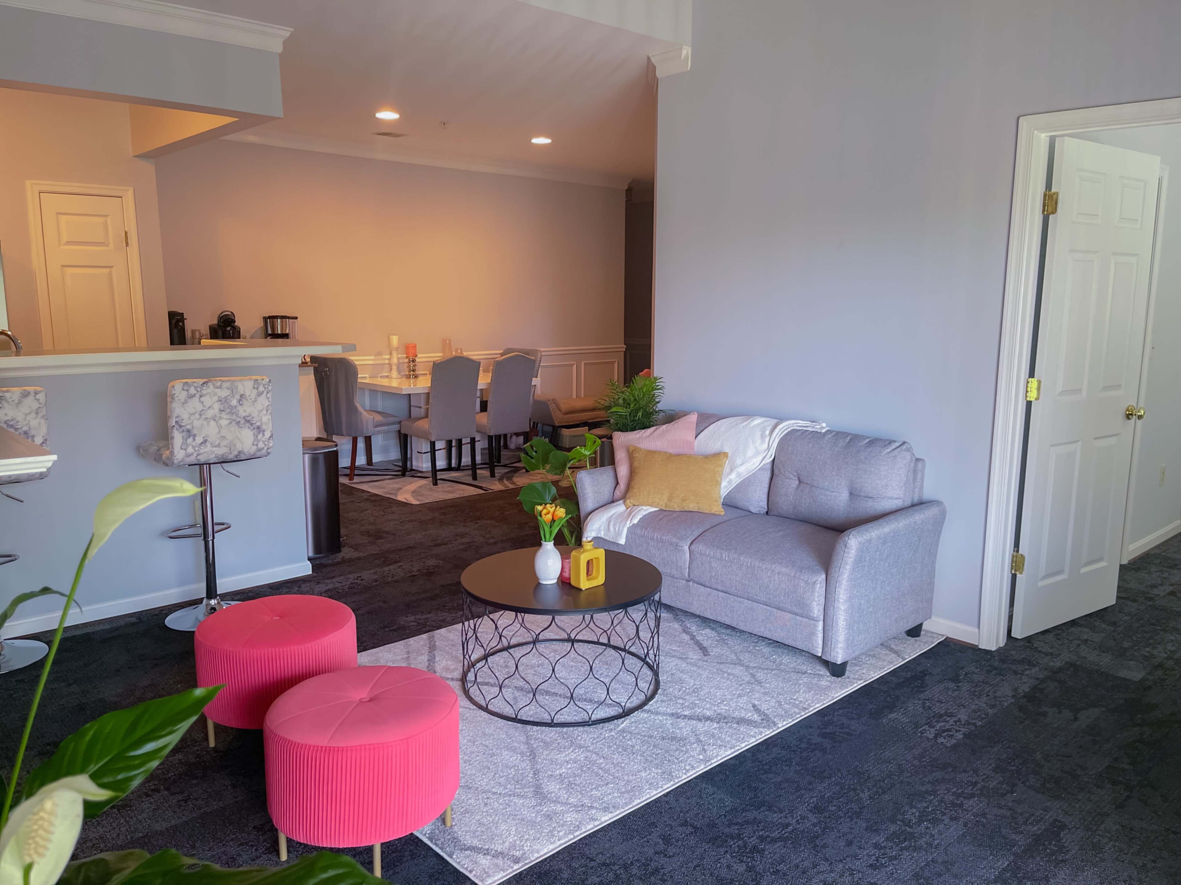 A modern living area features a gray couch, two pink ottomans, and a round coffee table, with a dining area visible in the background.