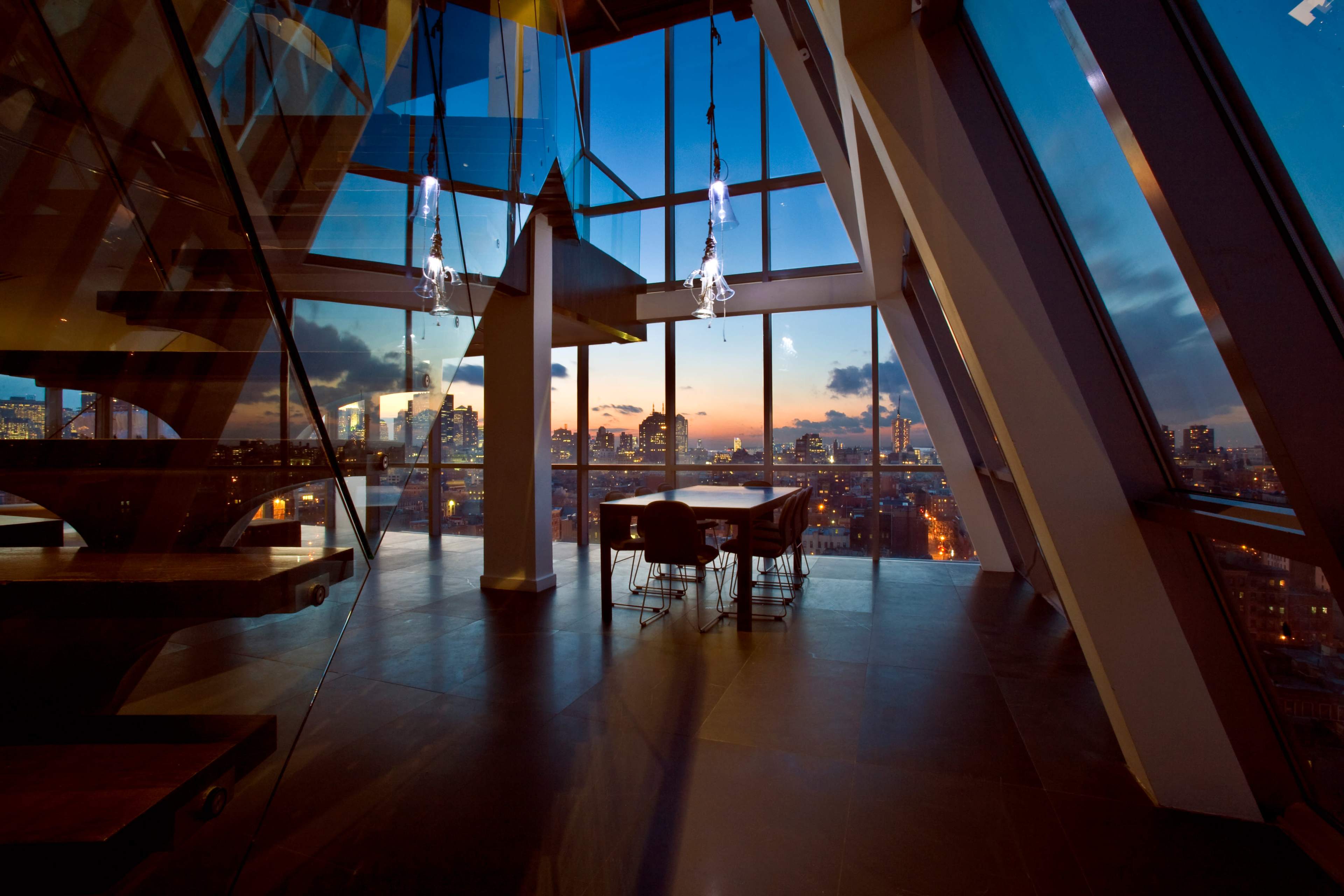 A modern interior features a staircase and a dining area with a large window showcasing a city skyline at sunset.